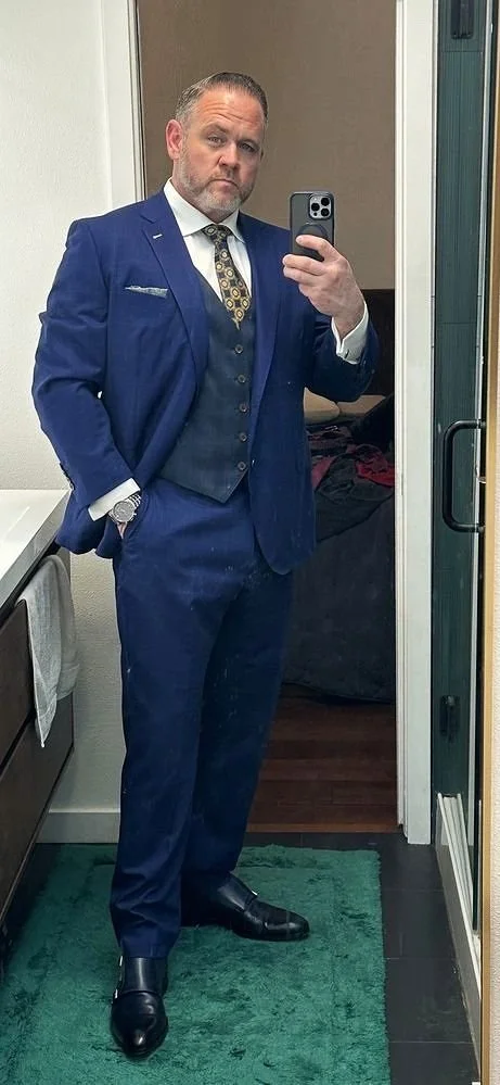 A man taking a mirror selfie in a dressing room, dressed in a blue suit with a white shirt and patterned tie, wearing black dress shoes and a watch.