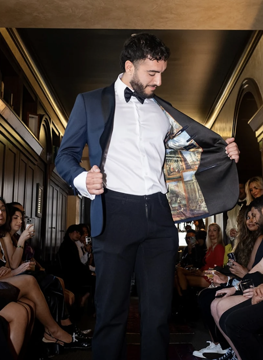 Man in formal tuxedo with a bowtie, holding open his tuxedo jacket to show a painted interior, standing in a room with people seated and taking photos.