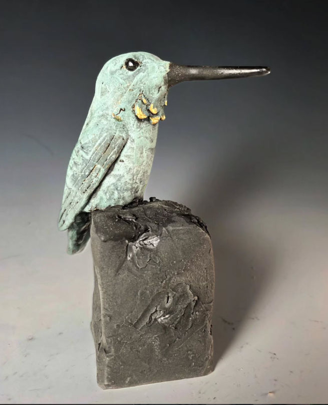 Hand built ceramic hummingbird sculpture in verdigris green with gold luster throat, perched on a rough stoneware block. A female Rufous, by the throat spots. The kind you see on the Eastern Slopes in July, holding a willow tip between feeding runs.
