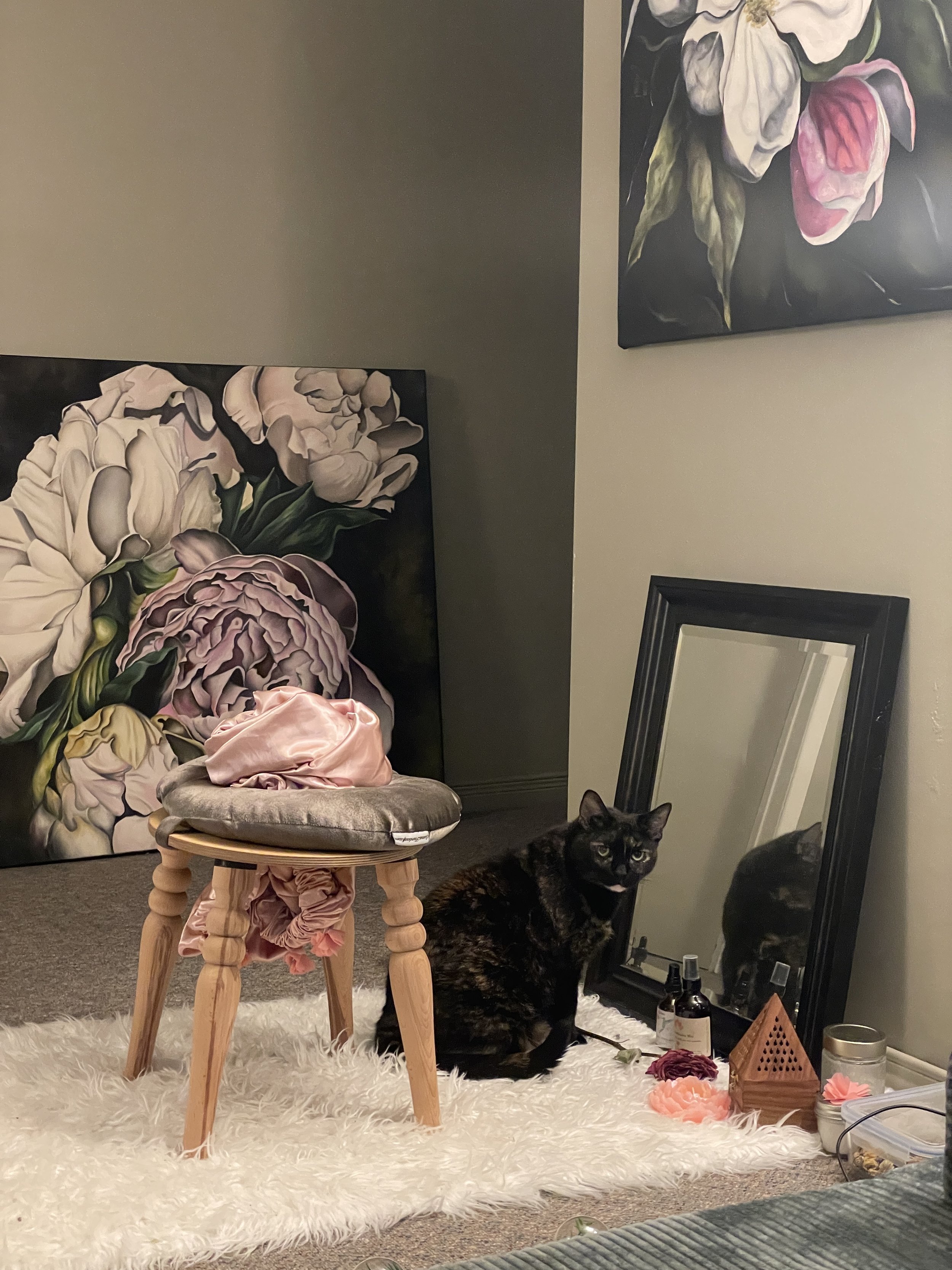 Interior scene with a black cat sitting on a white fluffy rug. Behind the rug, there is a small shiny stool, a large flower painting on the wall, a mirror leaning against the wall, a floor lamp with multiple bulbs, and some decorative candles on the floor. Pink artificial flowers are on the rug near the mirror.