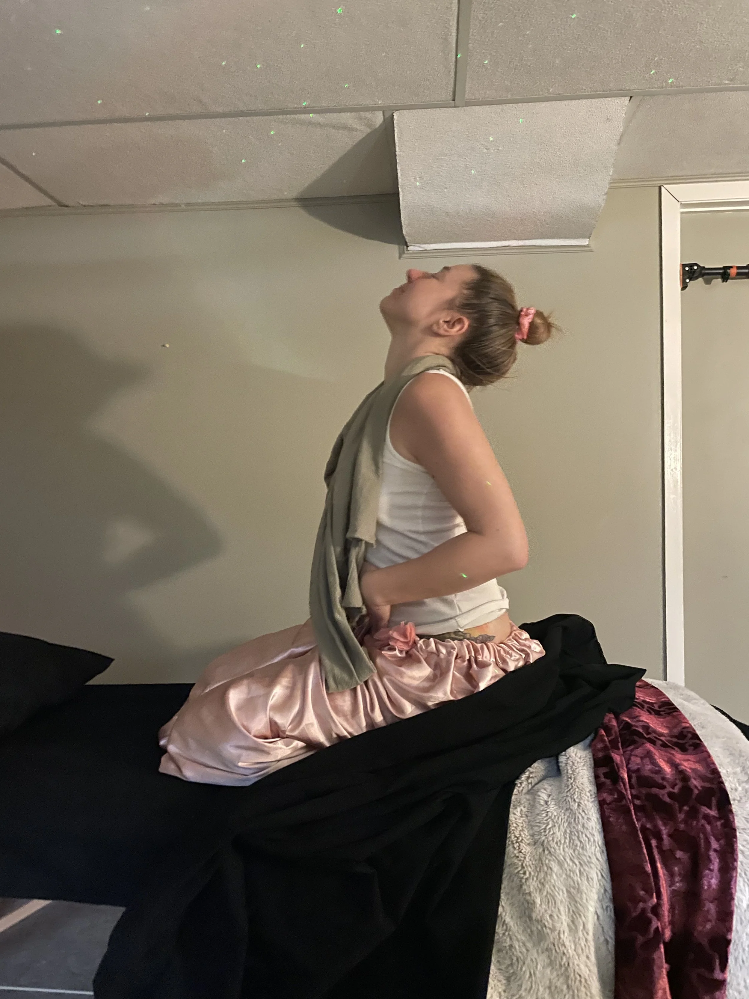 Interior scene with a black cat sitting on a white fluffy rug. Behind the rug, there is a small shiny stool, a large flower painting on the wall, a mirror leaning against the wall, a floor lamp with multiple bulbs, and some decorative candles on the floor. Pink artificial flowers are on the rug near the mirror.