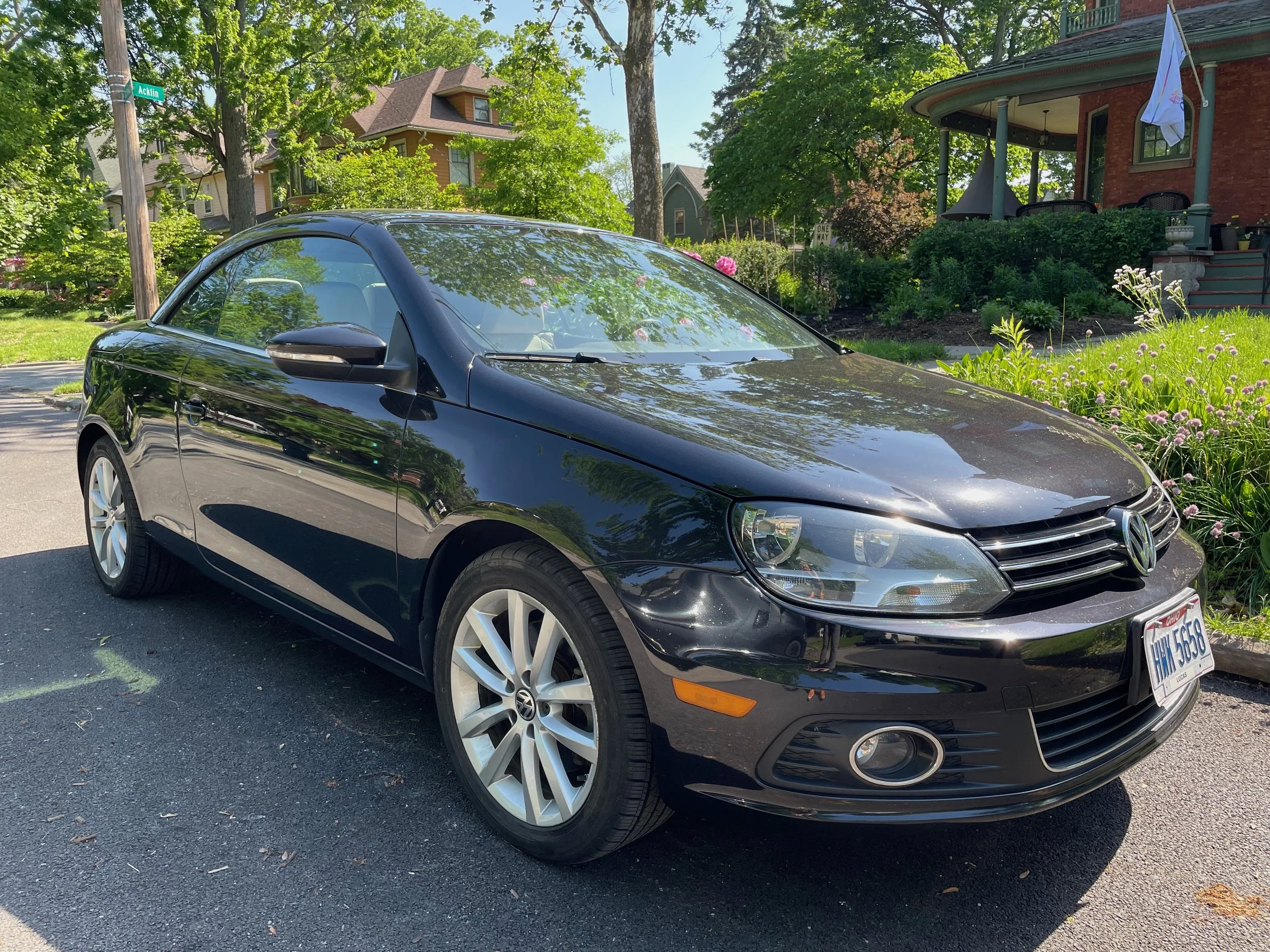 Black Volkswagen convertible car parked on the street in front of a house with a garden, trees, and a porch with chairs, under sunny weather.