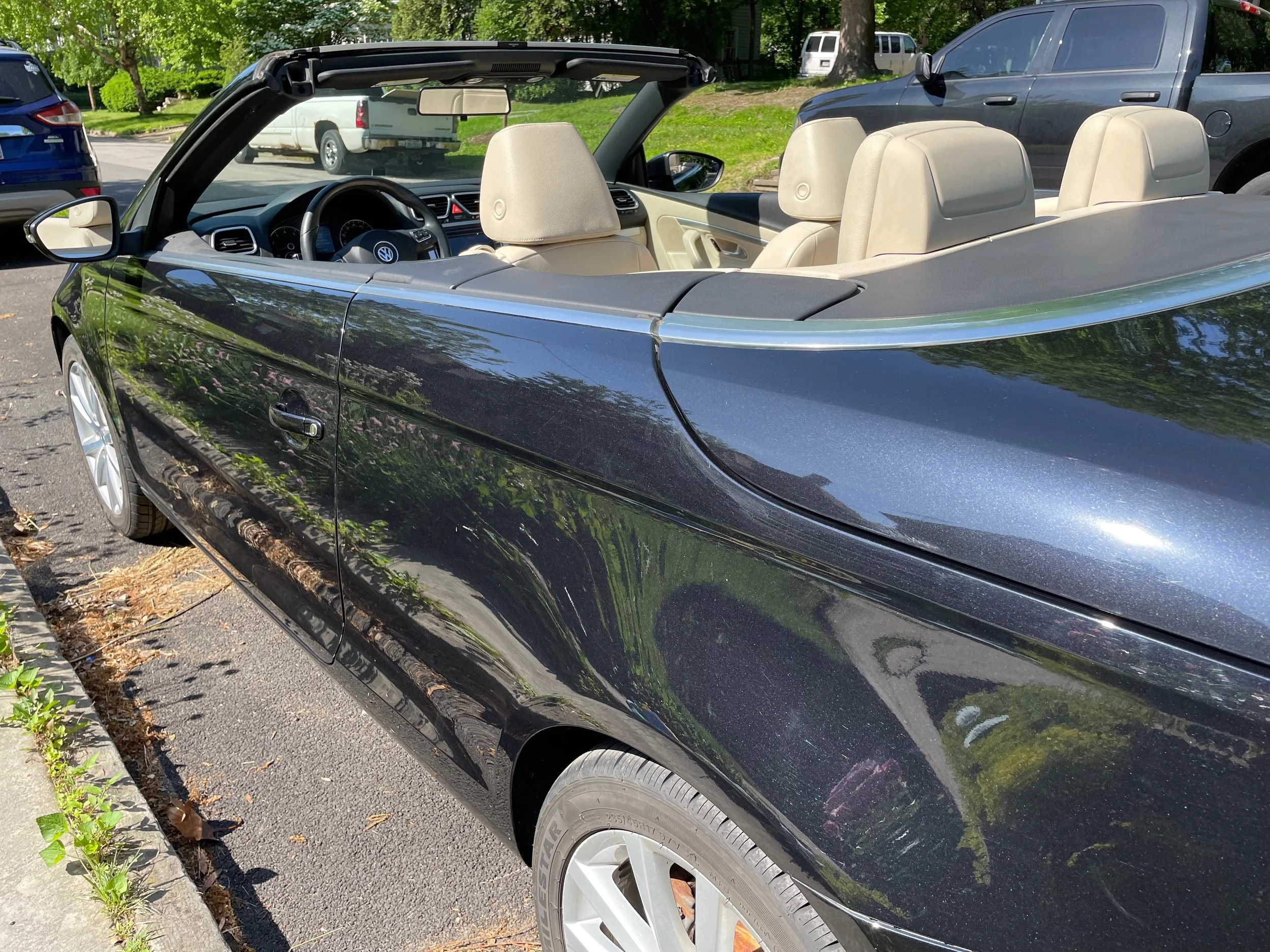 Black convertible car with beige interior parked on the street, with trees and other vehicles in the background.