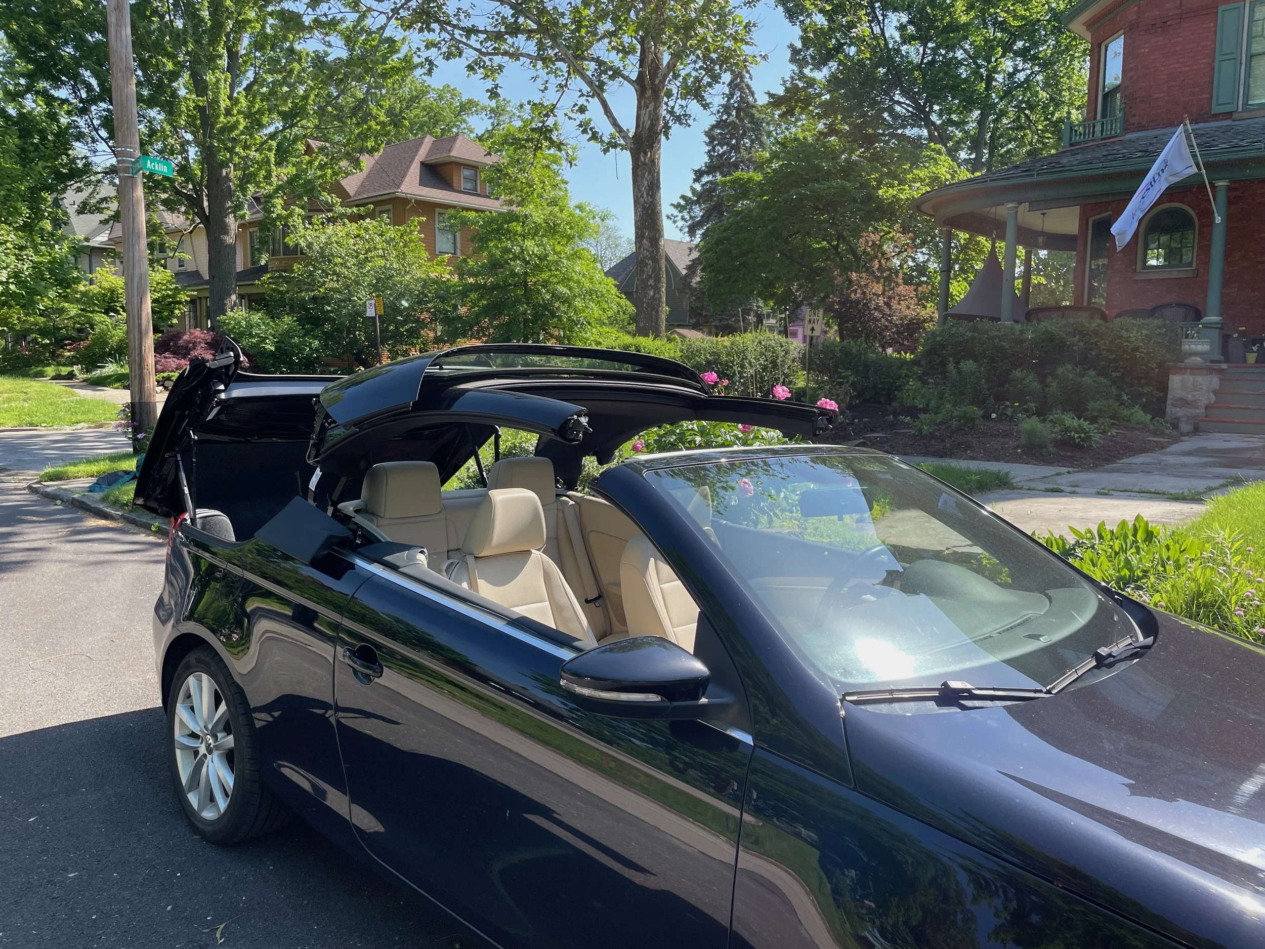 Black convertible car with beige interior parked on the street near a house with garden, trees, and a flag.