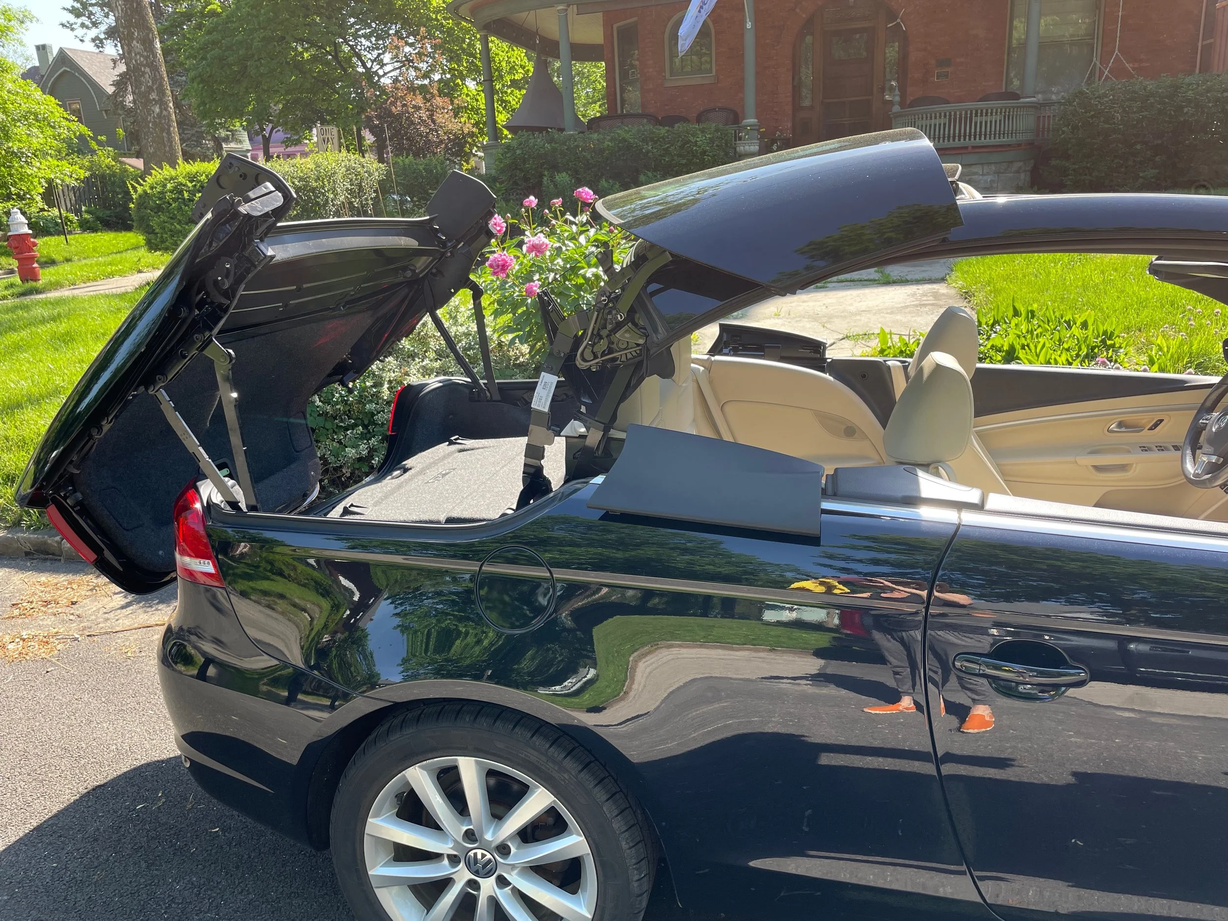 A black Volkswagen convertible car with its trunk and convertible roof open, parked on a residential street with green grass, trees, and houses in the background.