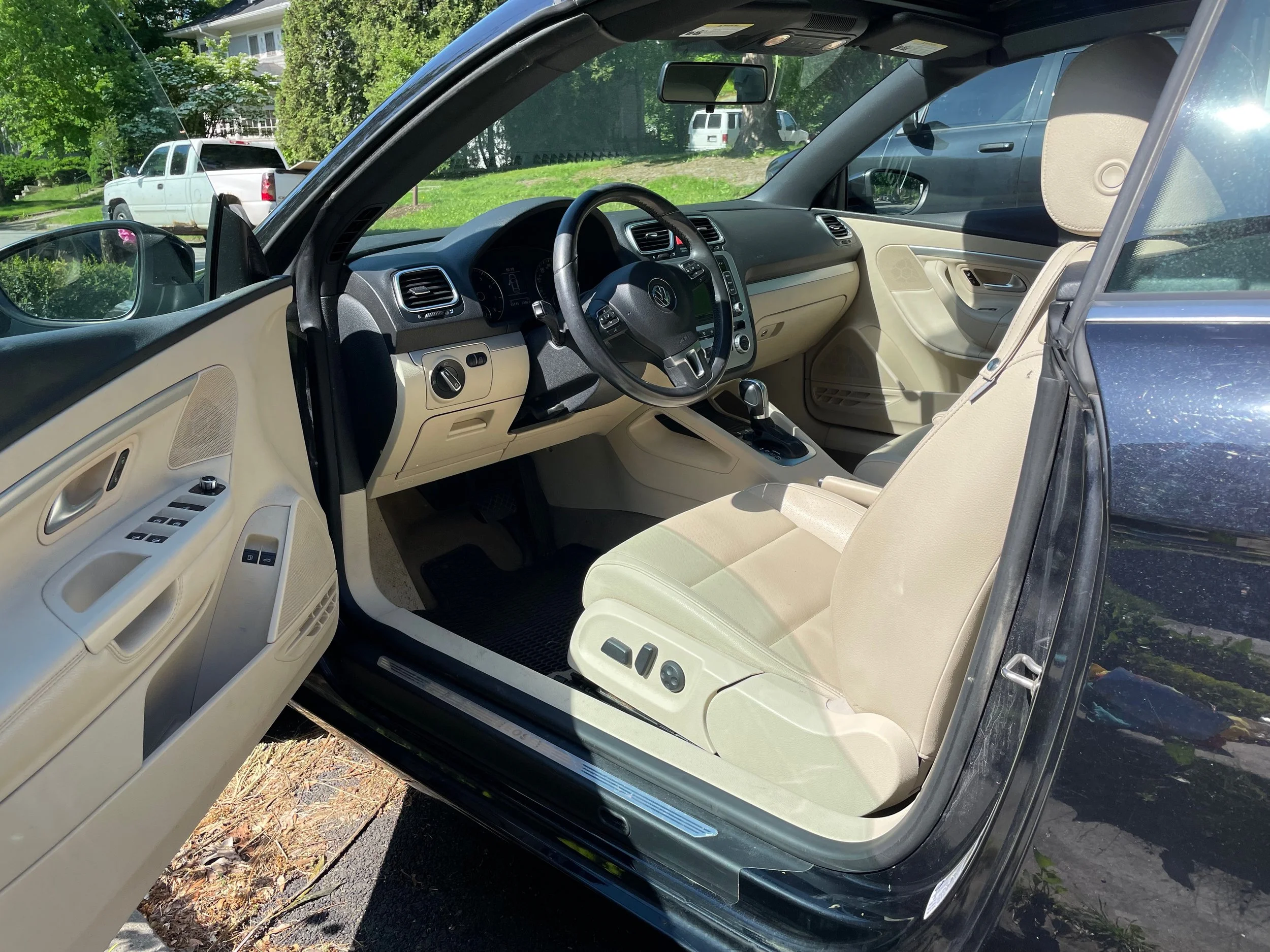 Inside view of a black vehicle with beige interior, showcasing the driver and front passenger seats, dashboard, and steering wheel. The doors are open, revealing controls on the door panels and the gear shift on the center console. External backgroun