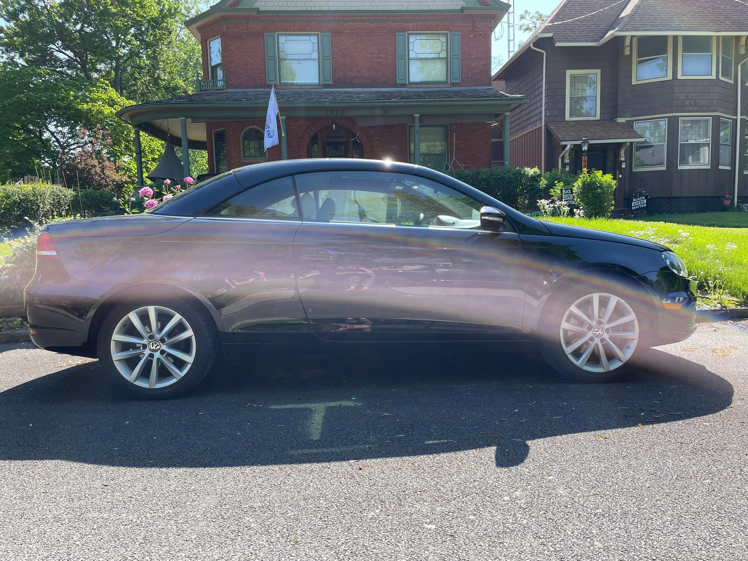 A black Volkswagen car parked on the street in front of houses with greenery and blooming flowers, under a partly sunny sky.