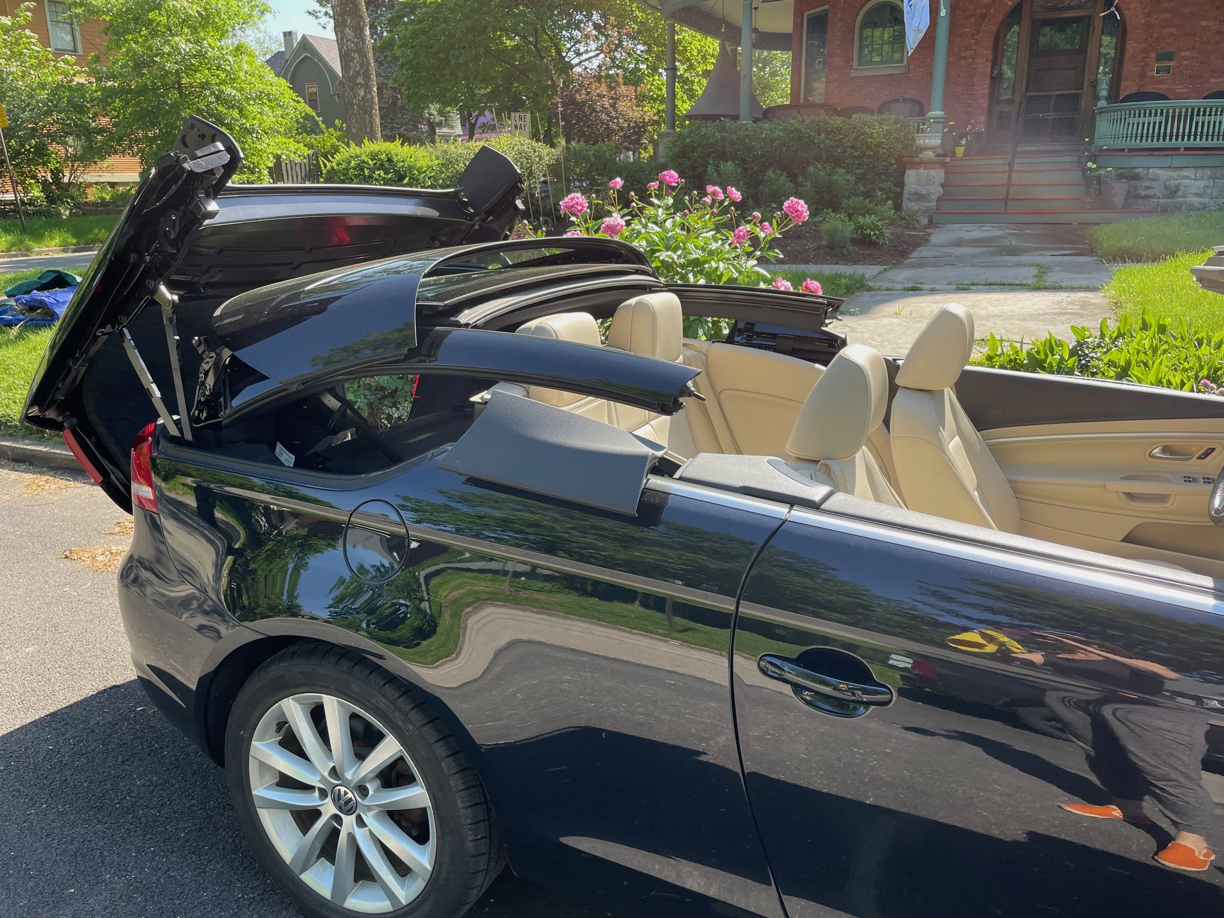 A black convertible car with a beige interior parked on a suburban street with open trunk, in front of a house with a garden and pink flowers.