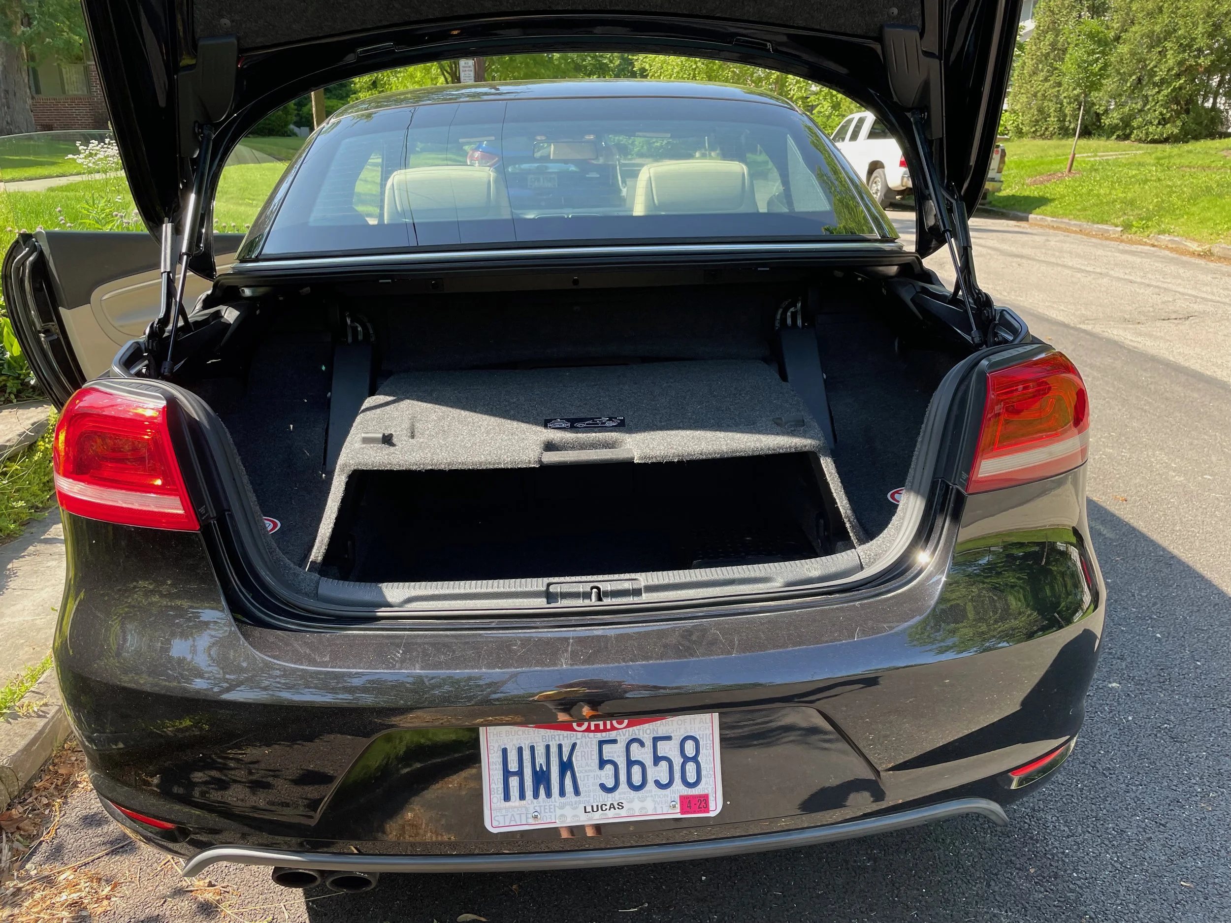 The back of a black sedan with the trunk open, revealing the cargo space. The car has Ohio license plates with the characters 'HWK 5658' and is parked on a suburban street with green grass and trees in the background.