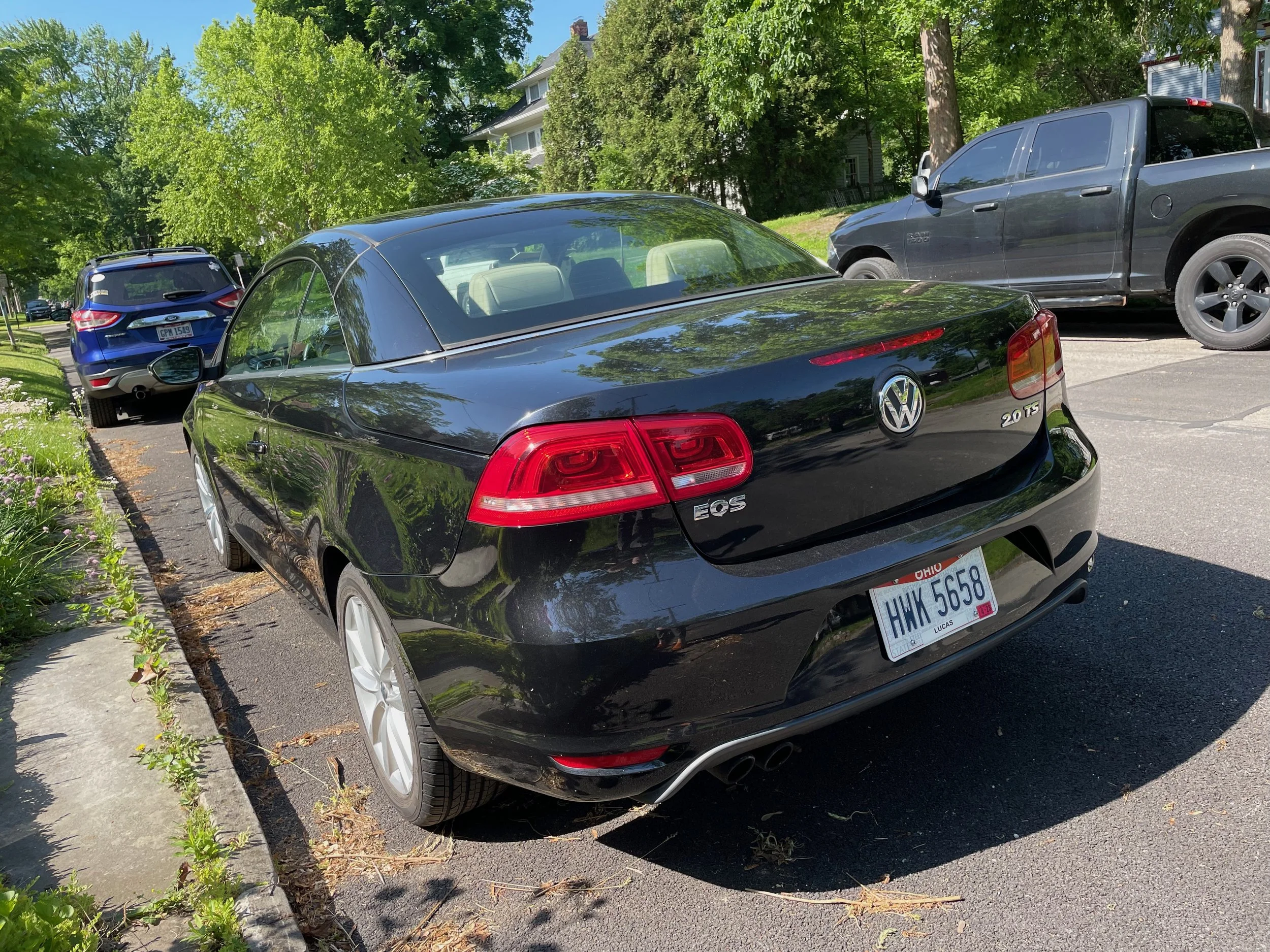 Black Volkswagen EOS convertible parked on the street with a beige interior, surrounded by other vehicles and green trees.