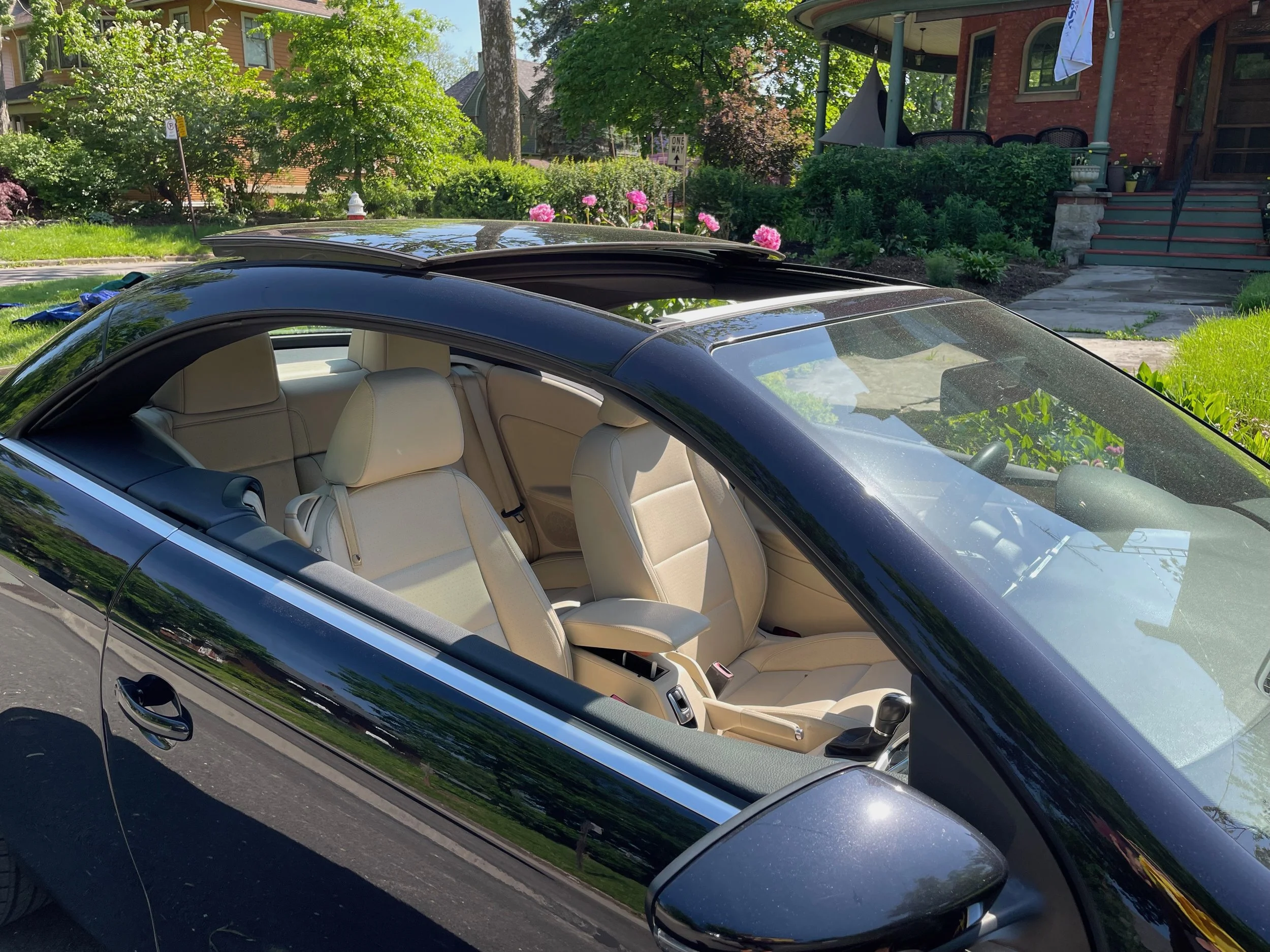 A black convertible car with a beige interior parked in a residential neighborhood with a house, garden, and blooming flowers in the background.