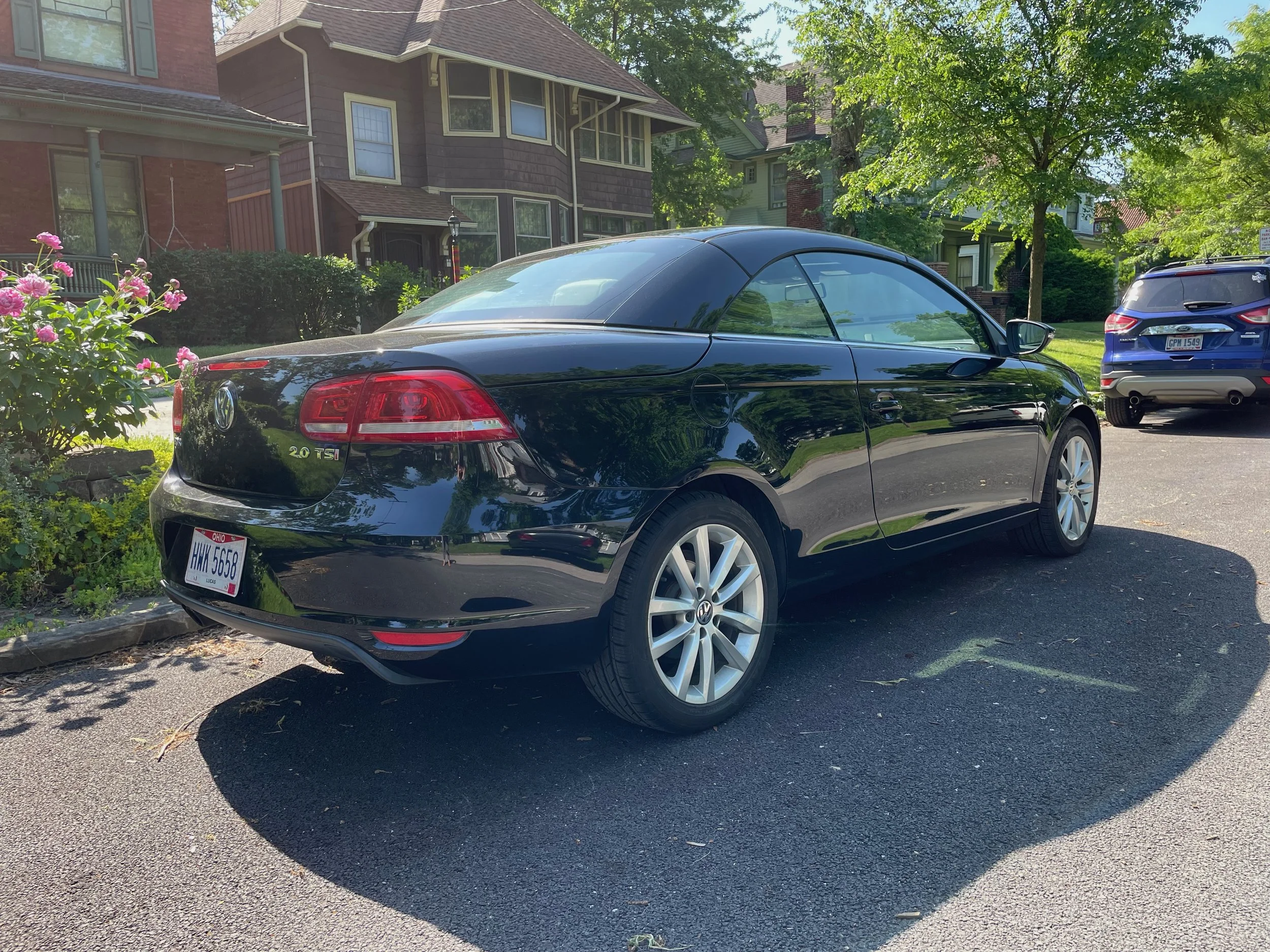 Black Volkswagen convertible car parked on a residential street with houses and trees in the background.
