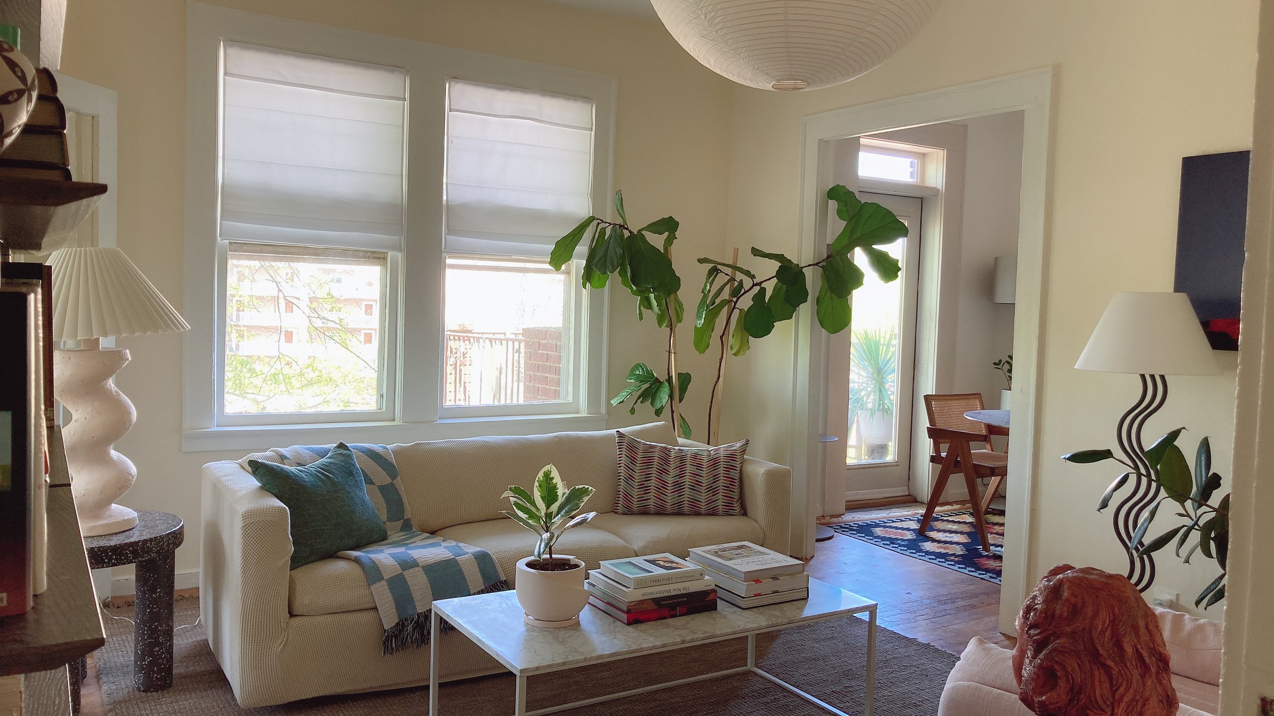 Living room with beige couch, throw pillows, and a coffee table with books and a plant. Large windows with blinds, a tall plant near the window, and a doorway leading to another room with a rug and a small table with chairs. Various lamps and plants 