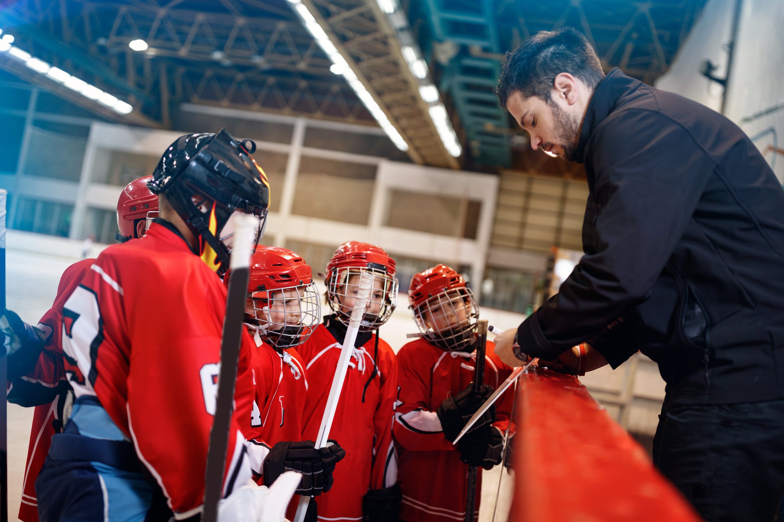 A youth hockey coach giving instructions to a team of young players dressed in red jerseys and helmets on an indoor ice rink.