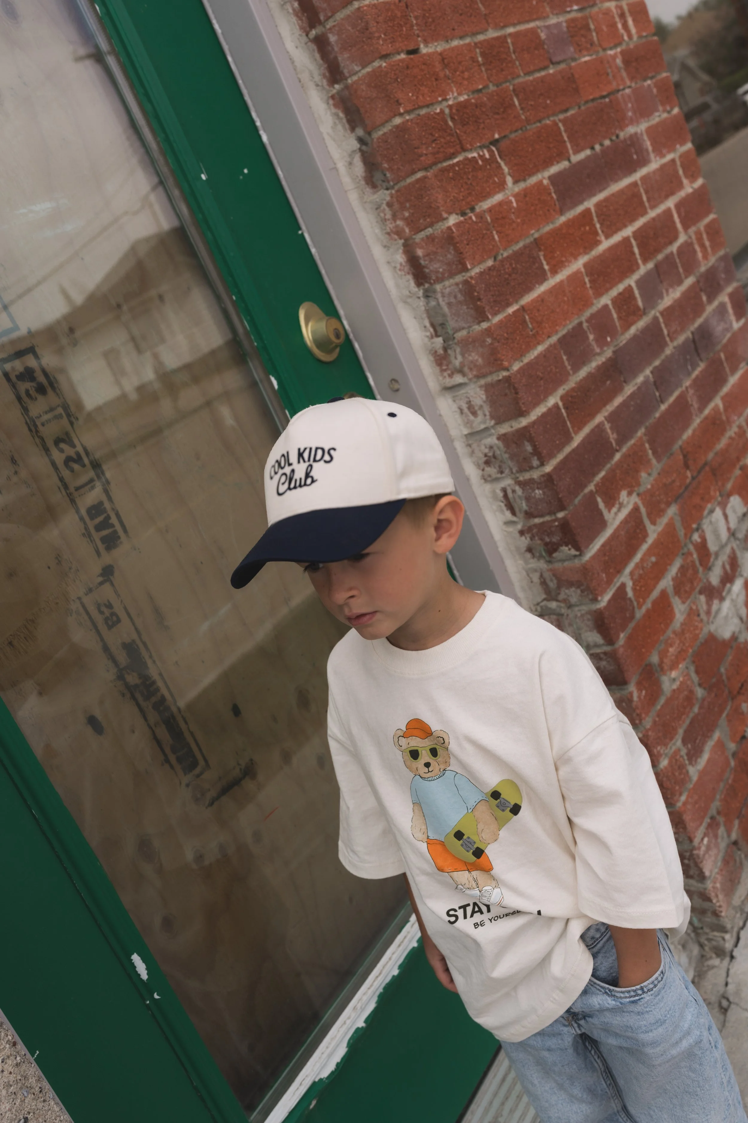 A young boy wearing a white baseball cap with the words 'Cool Kids Club' and a graphic of a bear in sunglasses, white t-shirt with a bear graphic, and blue jeans, standing in front of a glass door with a brick wall behind him.
