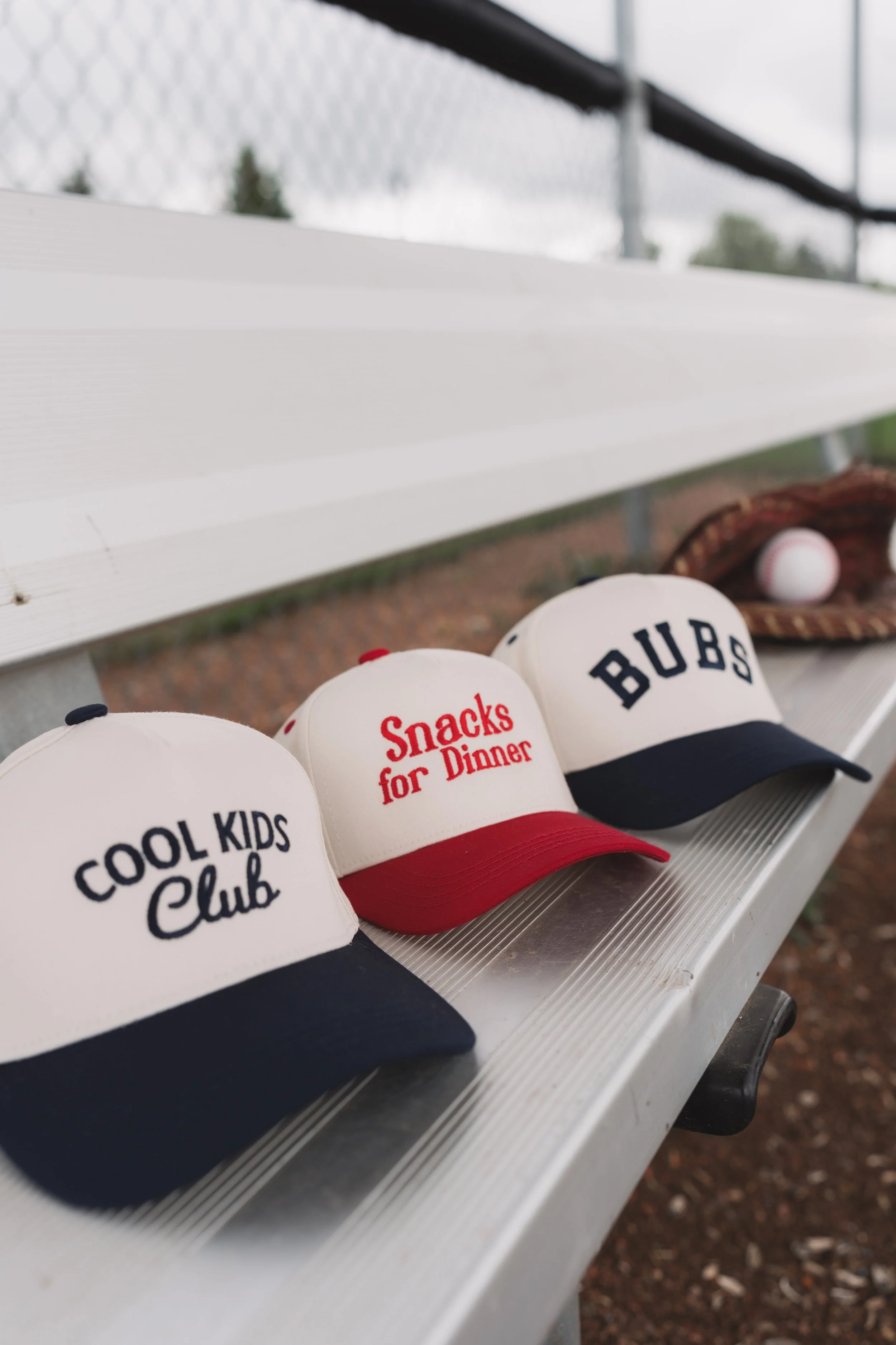 A row of three baseball-style caps on a bench, with slogans reading 'Cool Kids Club', 'Snacks for Dinner', and 'Bubs'. A baseball glove and ball are visible in the background.