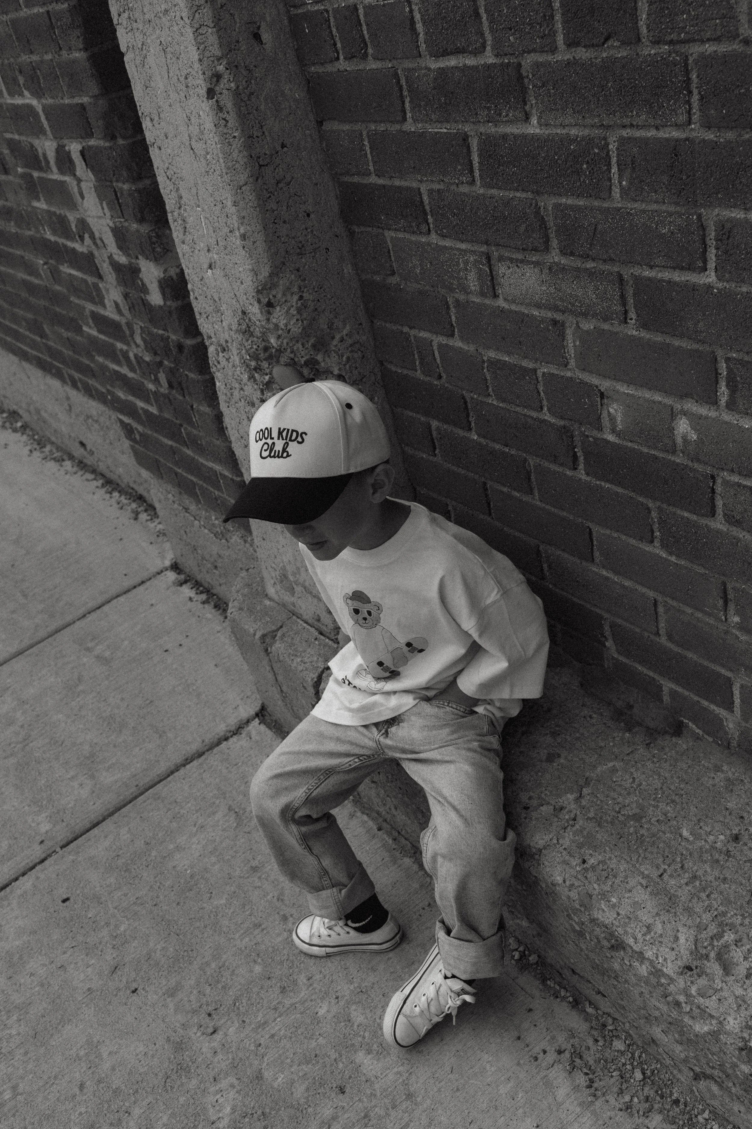 A young boy is sitting on the sidewalk leaning against a brick wall, wearing a baseball cap that says 'Cool Kids Club', a t-shirt with a teddy bear design, and sneakers.