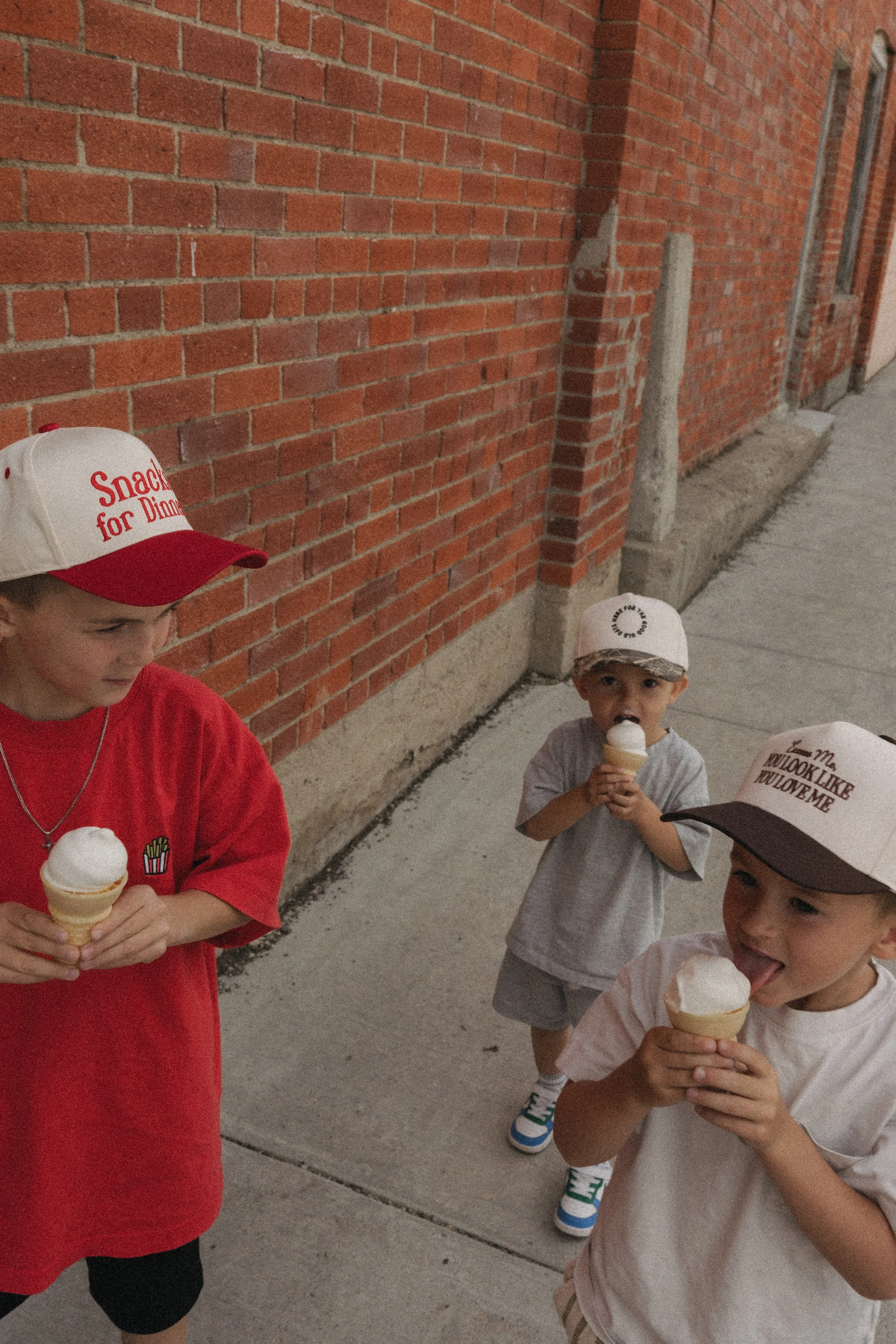 Three children enjoying ice cream cones outside against a brick wall. One boy in a red shirt and hat with text about snacks, a girl in a striped shirt, and another boy in a gray shirt, some wearing hats.