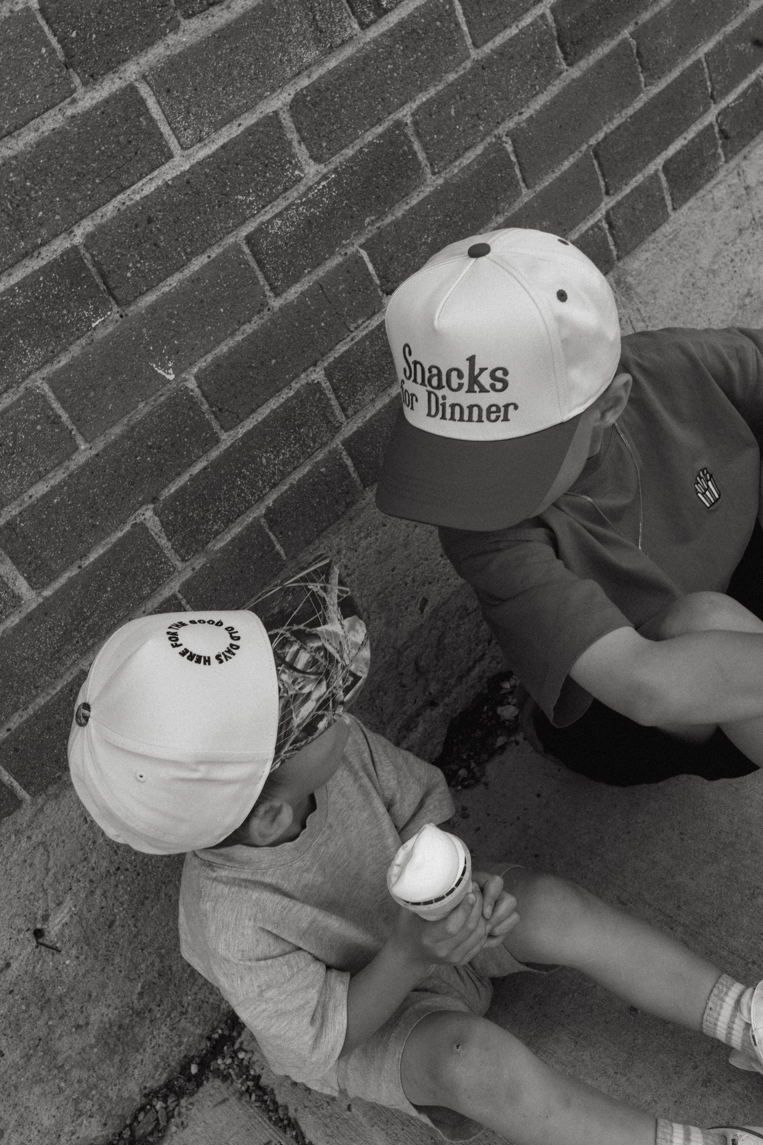 Two children sitting against a brick wall, wearing hats and casual clothing. One child is holding a handheld fan and the other is holding a paper cup. They are outdoors on a sidewalk.