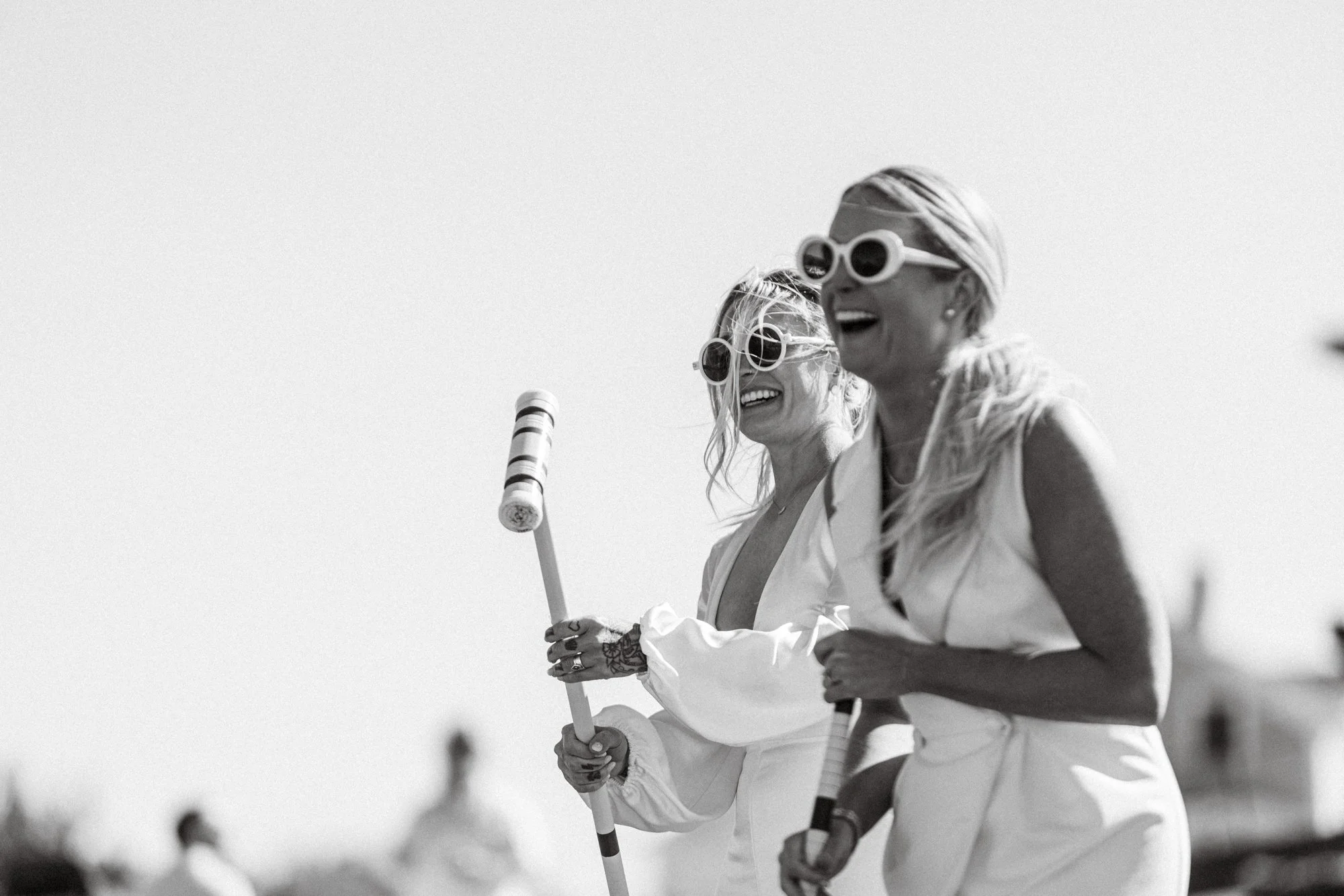 Two women wearing large sunglasses and white outfits, holding a croquet mallet, smiling, outdoors in bright sunlight.