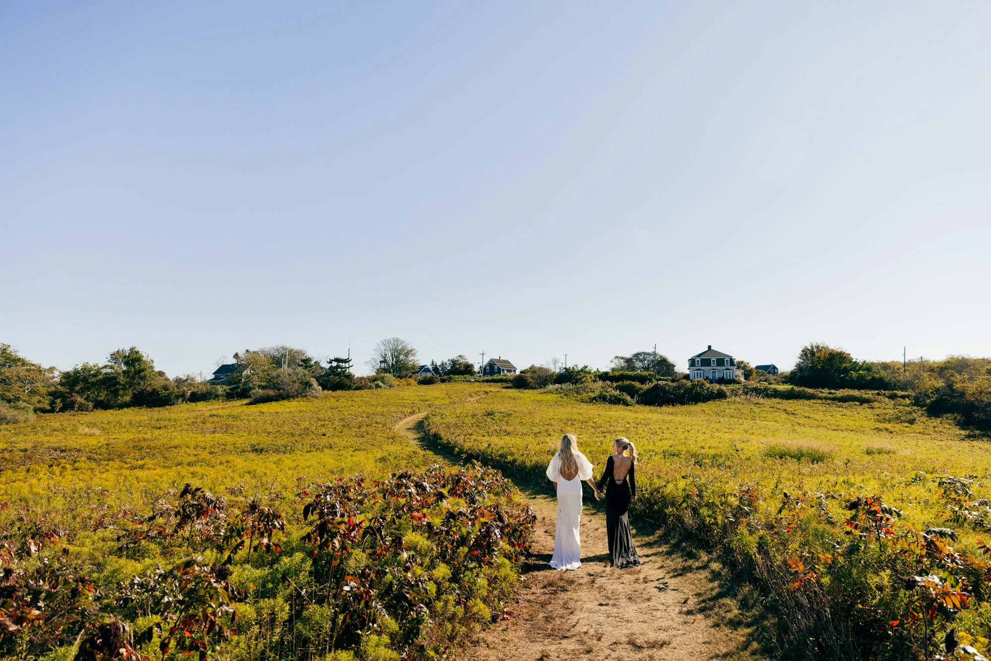 Two women in dresses walking hand in hand down a dirt path through a sunny yellow-green field with houses in the distance.