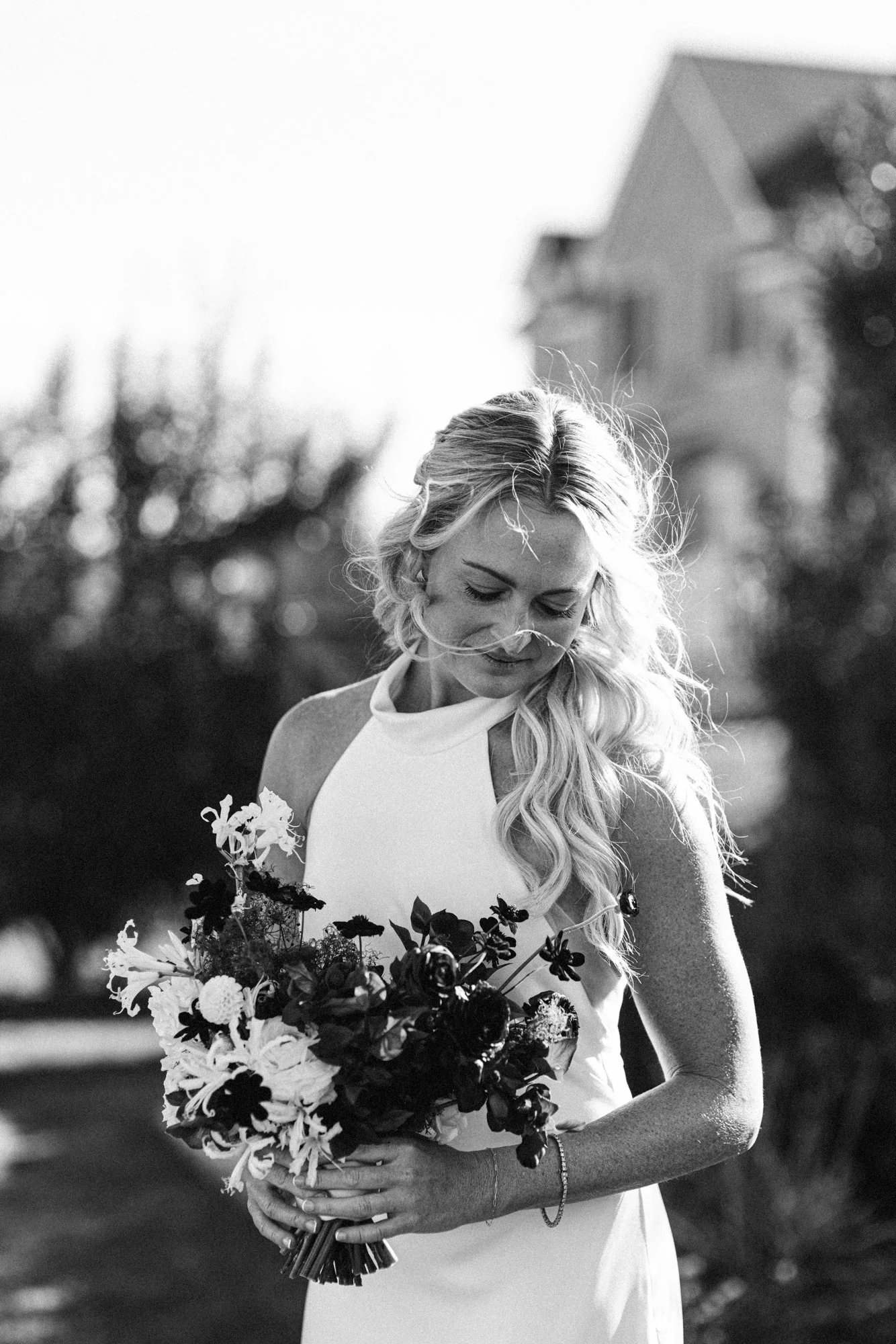 Black and white photo of a woman in a white dress holding a bouquet of flowers, outdoors with a building in the background.