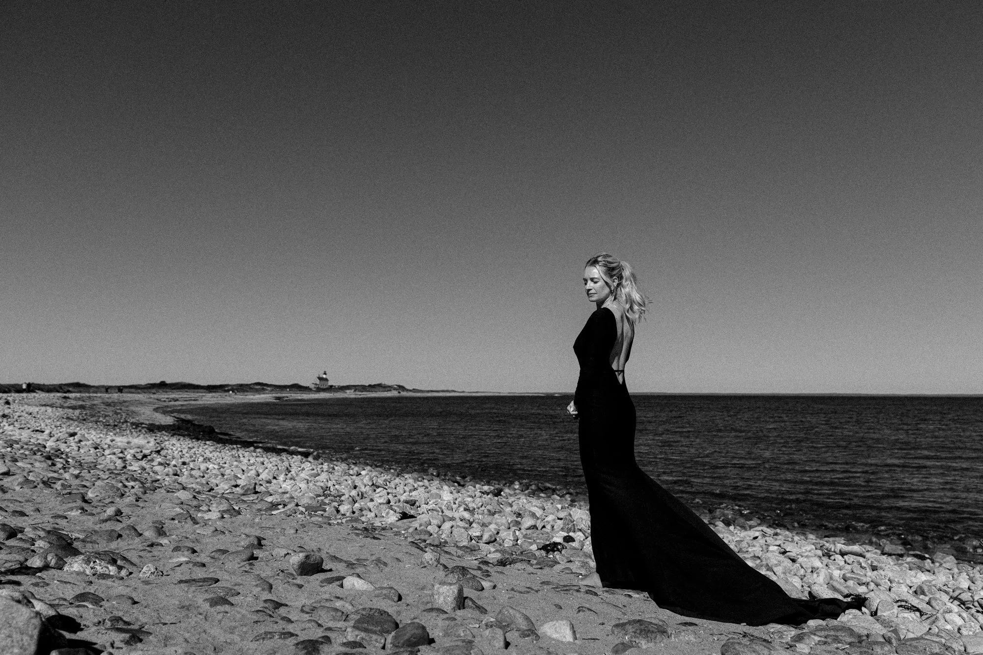 A woman in a long black dress standing on a rocky beach near the ocean with a lighthouse in the distance, black and white photograph.