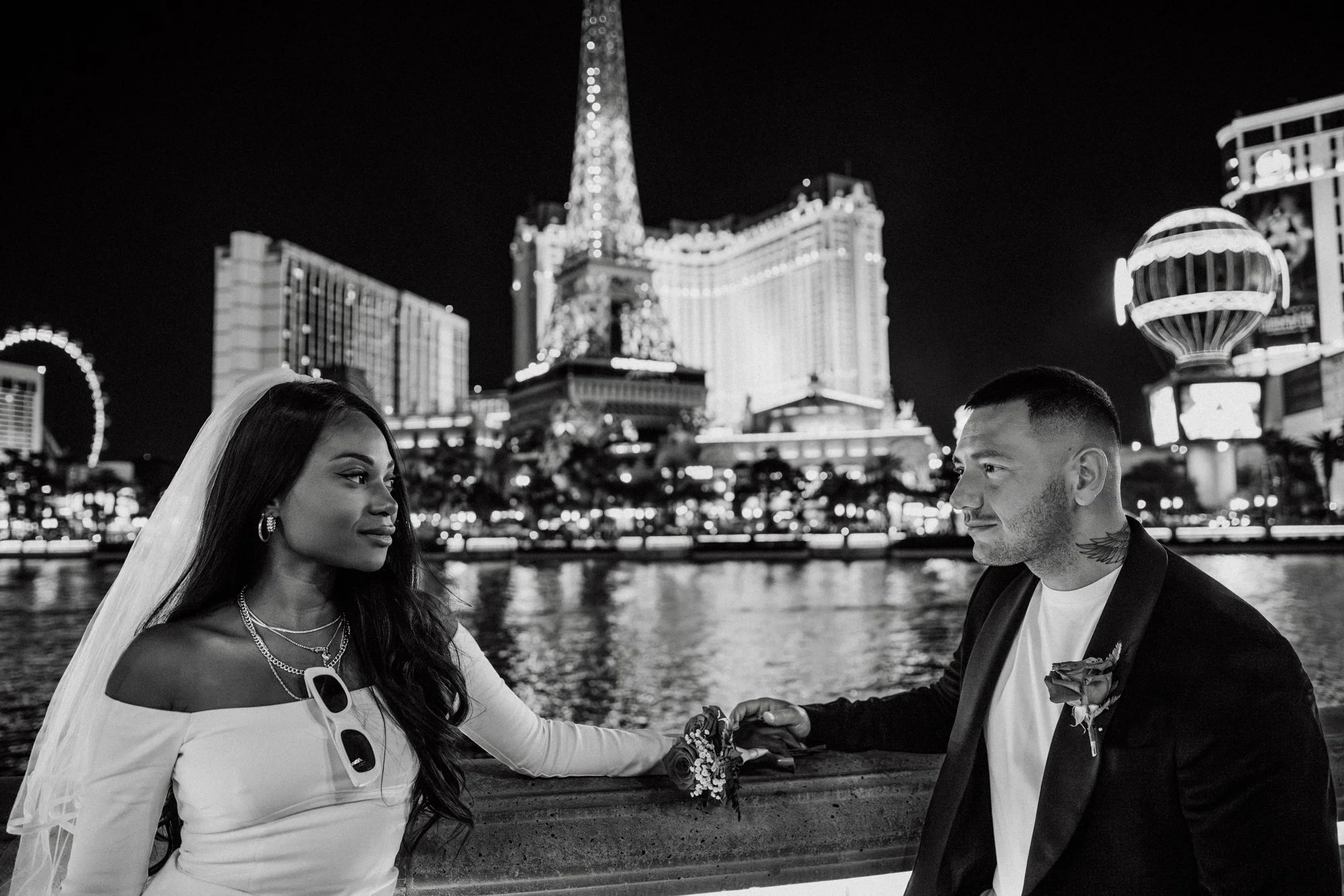 A black and white photo of a couple on their wedding night, holding hands and gazing at each other on a bridge by a river at night in Las Vegas, with city lights and the Las Vegas Strip's famous buildings behind them.