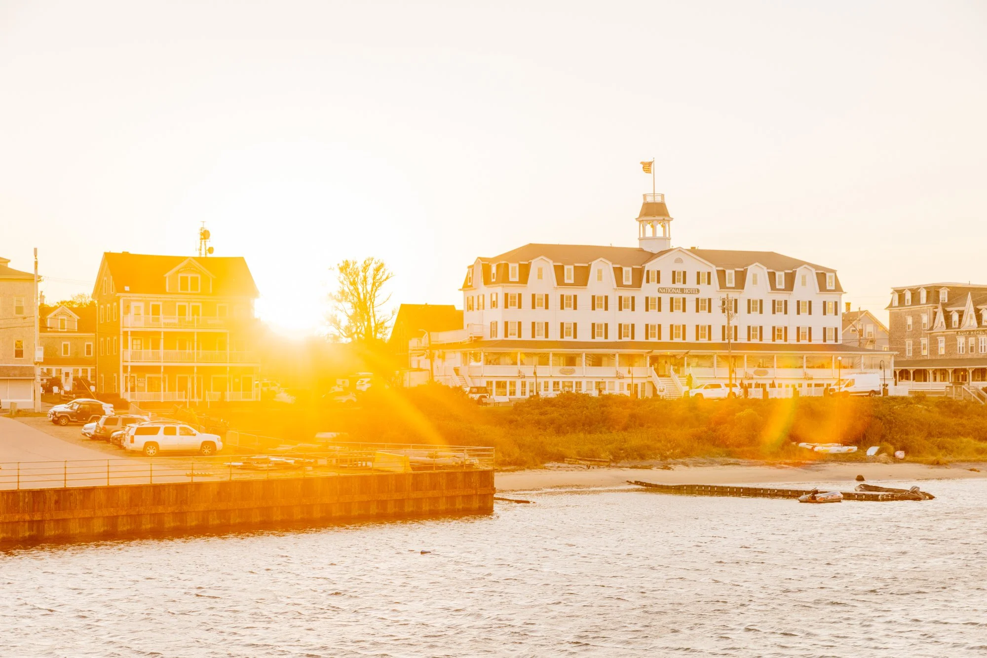 Sunset over a waterfront hotel and surrounding buildings with parked cars and a dock in the foreground.
