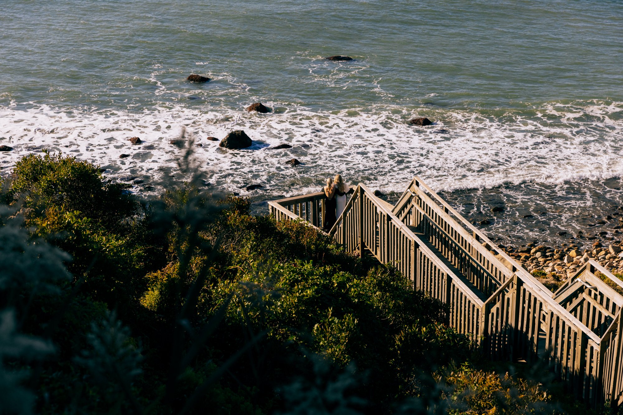 Two people standing on a wooden staircase overlooking the beach and ocean waves.