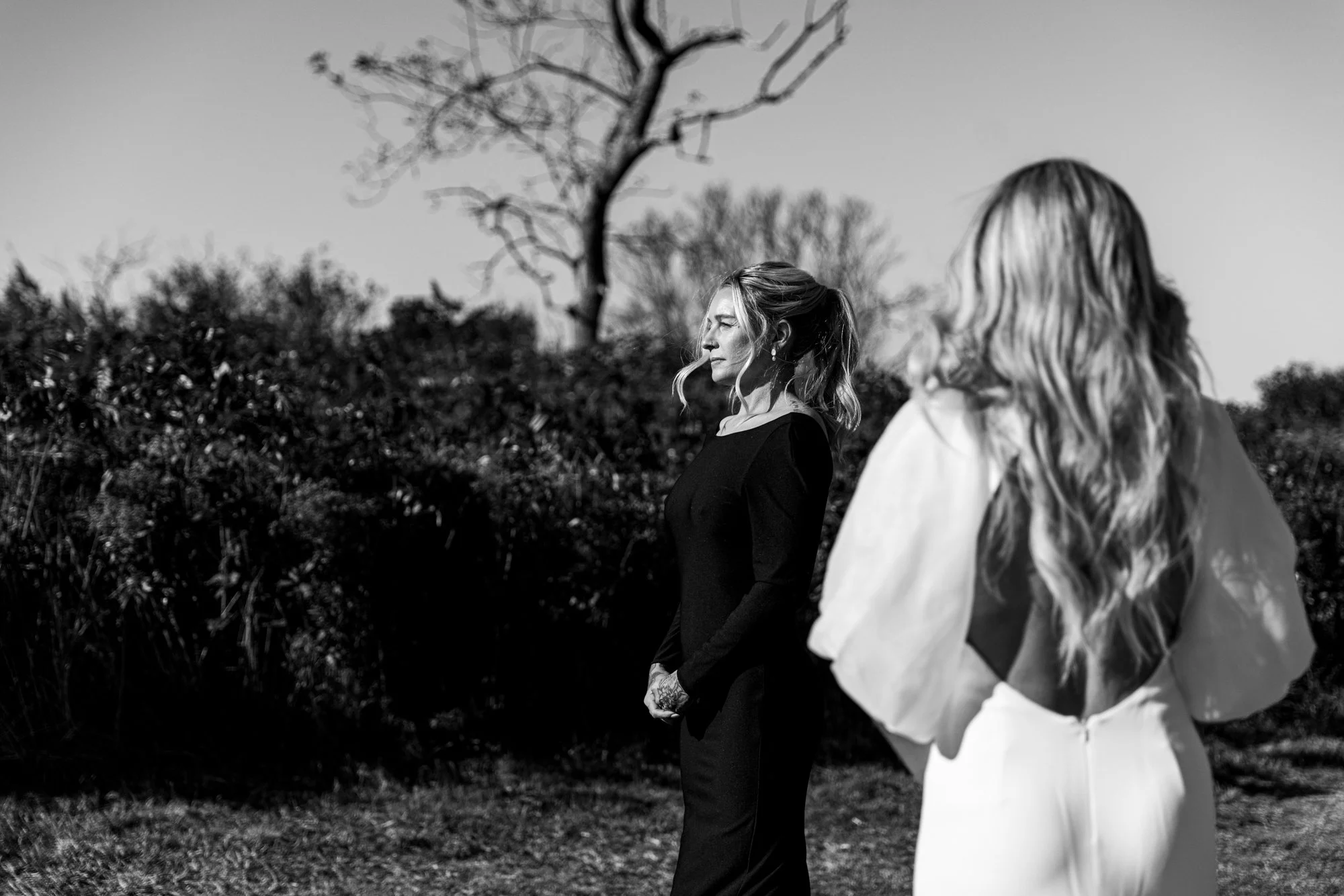 Black and white photo of two women standing outdoors with bushes and a leafless tree in the background. One woman is facing to the side with wavy hair tied back, wearing a dark dress. The other woman, with wavy hair and wearing a light-colored outfit