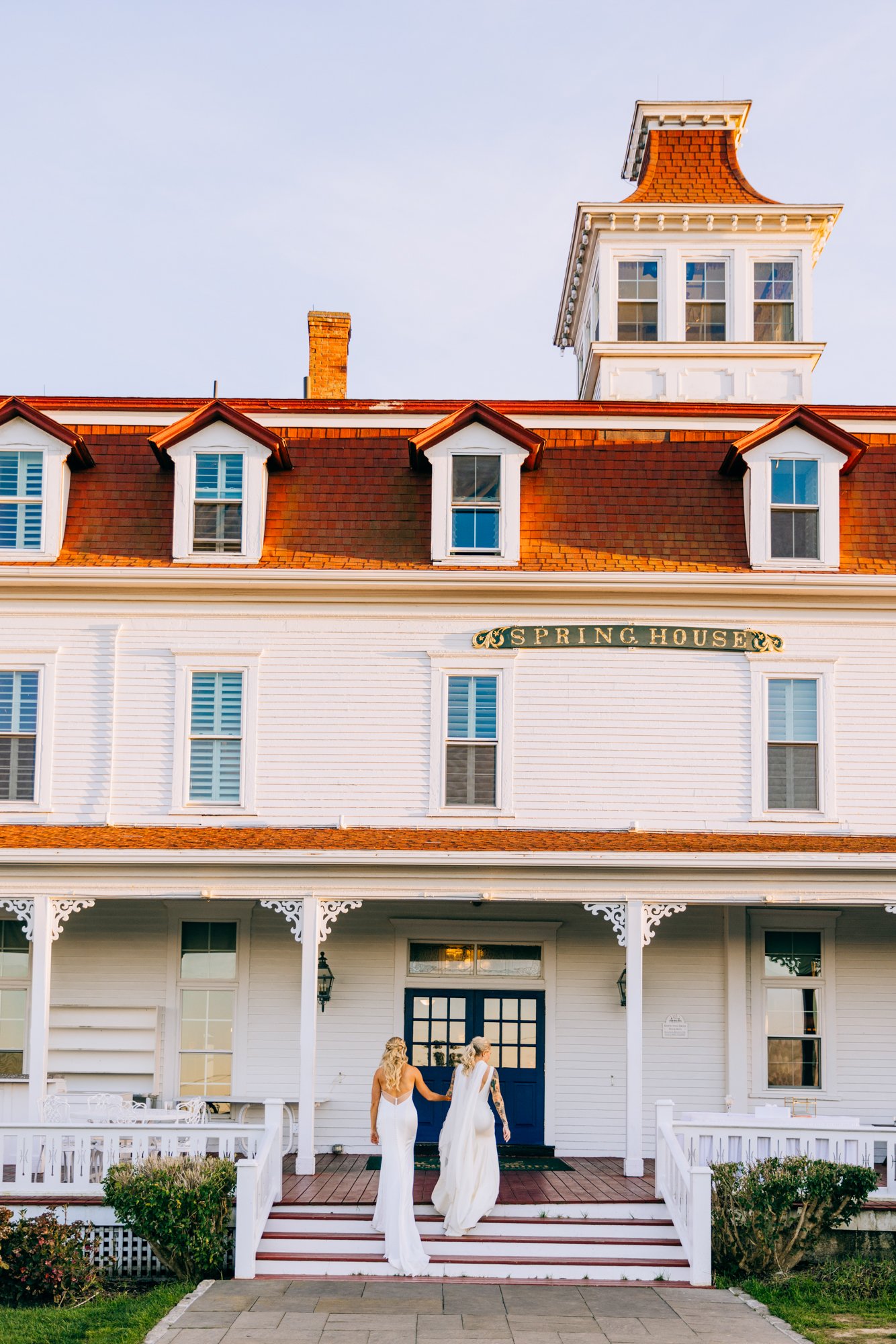 Two women in white dresses walking up the stairs of a large white building with a sign that says 'Spring House' on it, with a clear sky in the background.