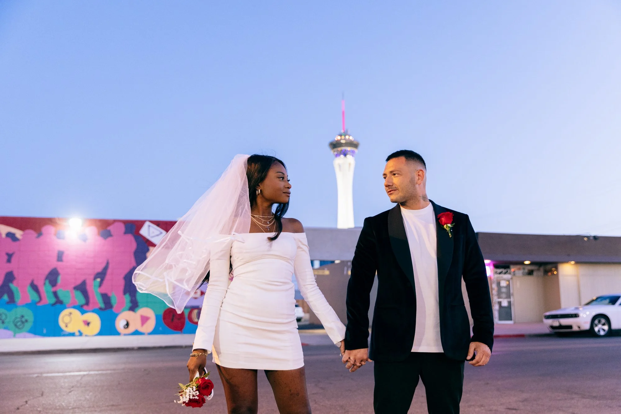 A bride and groom holding hands and looking at each other in front of a mural with the Stratosphere Tower in Las Vegas during dusk.