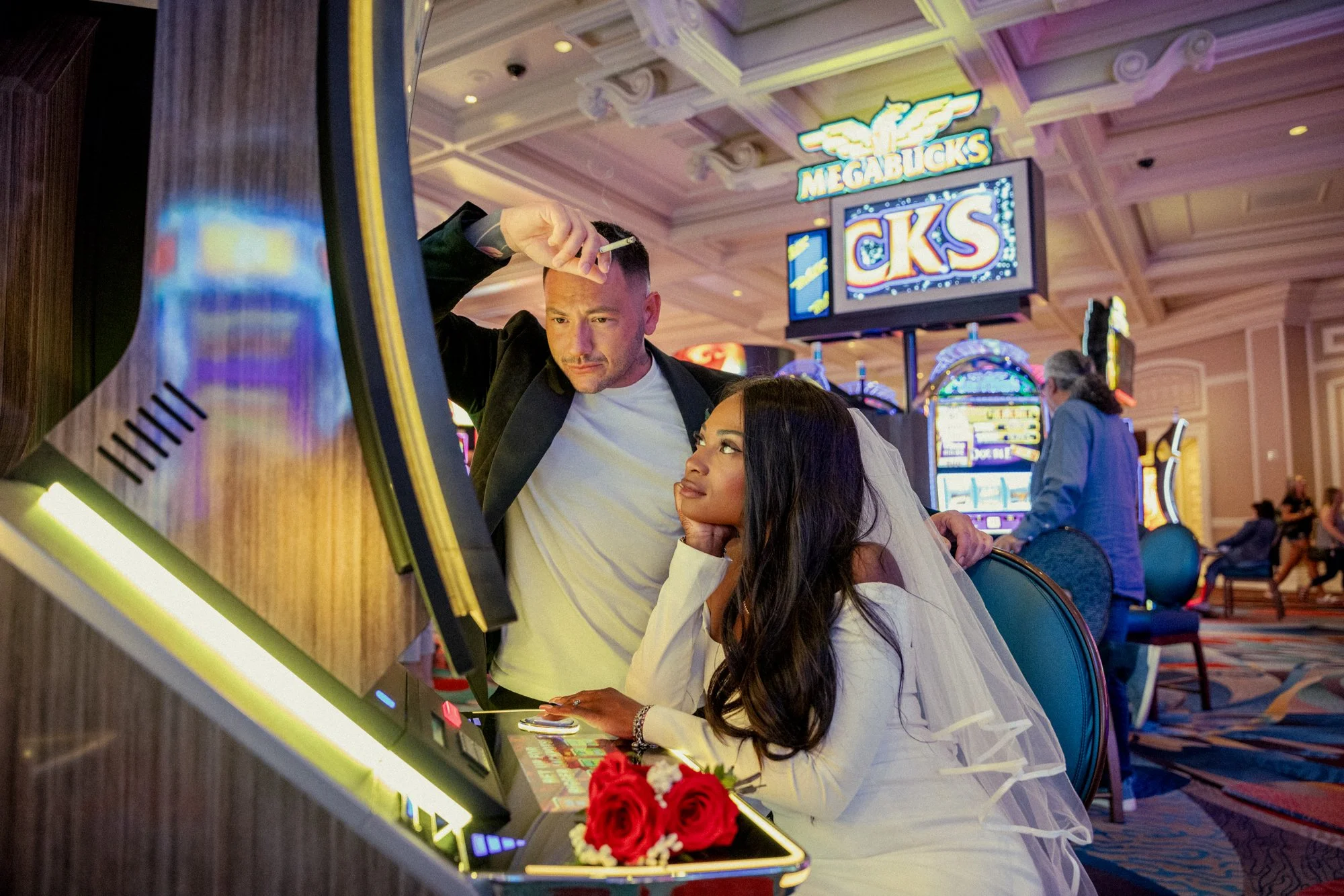 A man and woman at a slot machine in a casino. The woman is wearing a wedding veil and a white dress, holding a bouquet of red roses, while the man is dressed casually with a blazer. The casino features bright lights and multiple slot machines, with 
