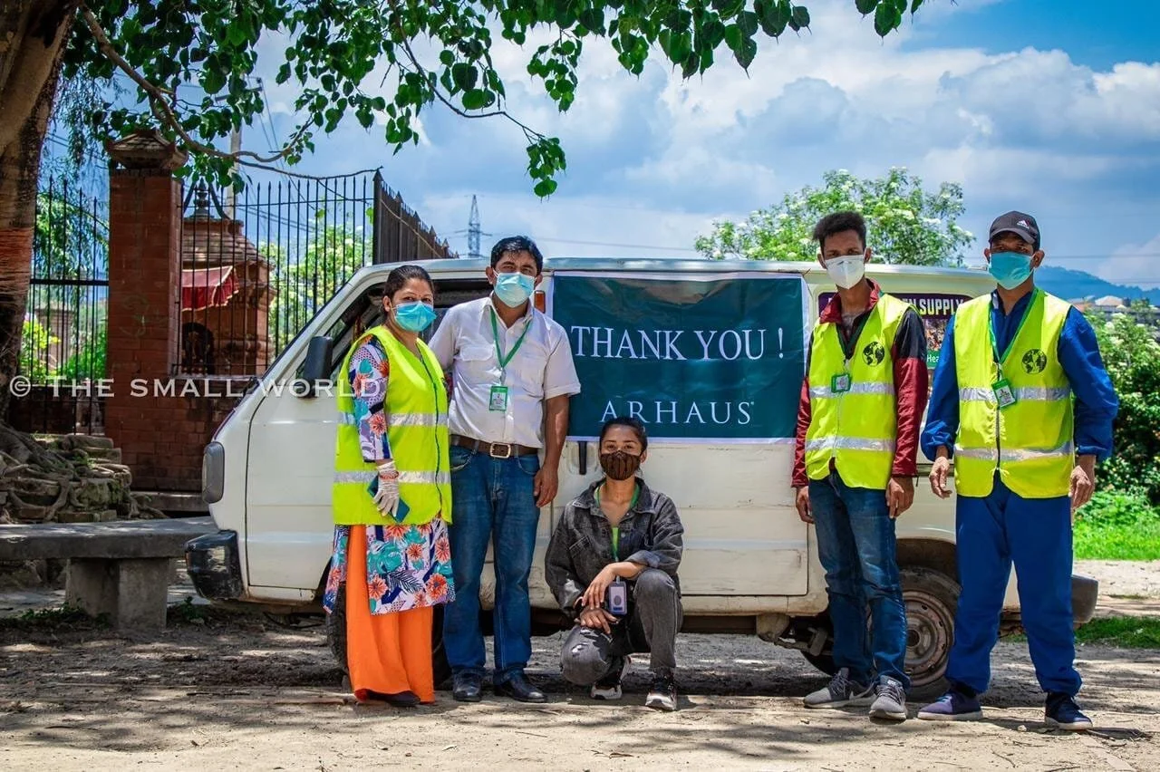 Group of five people wearing face masks and reflective vests standing in front of a small white vehicle with a blue banner that reads "Thank You! ARHAUS." They are outdoors with trees and cloudy sky in the background.