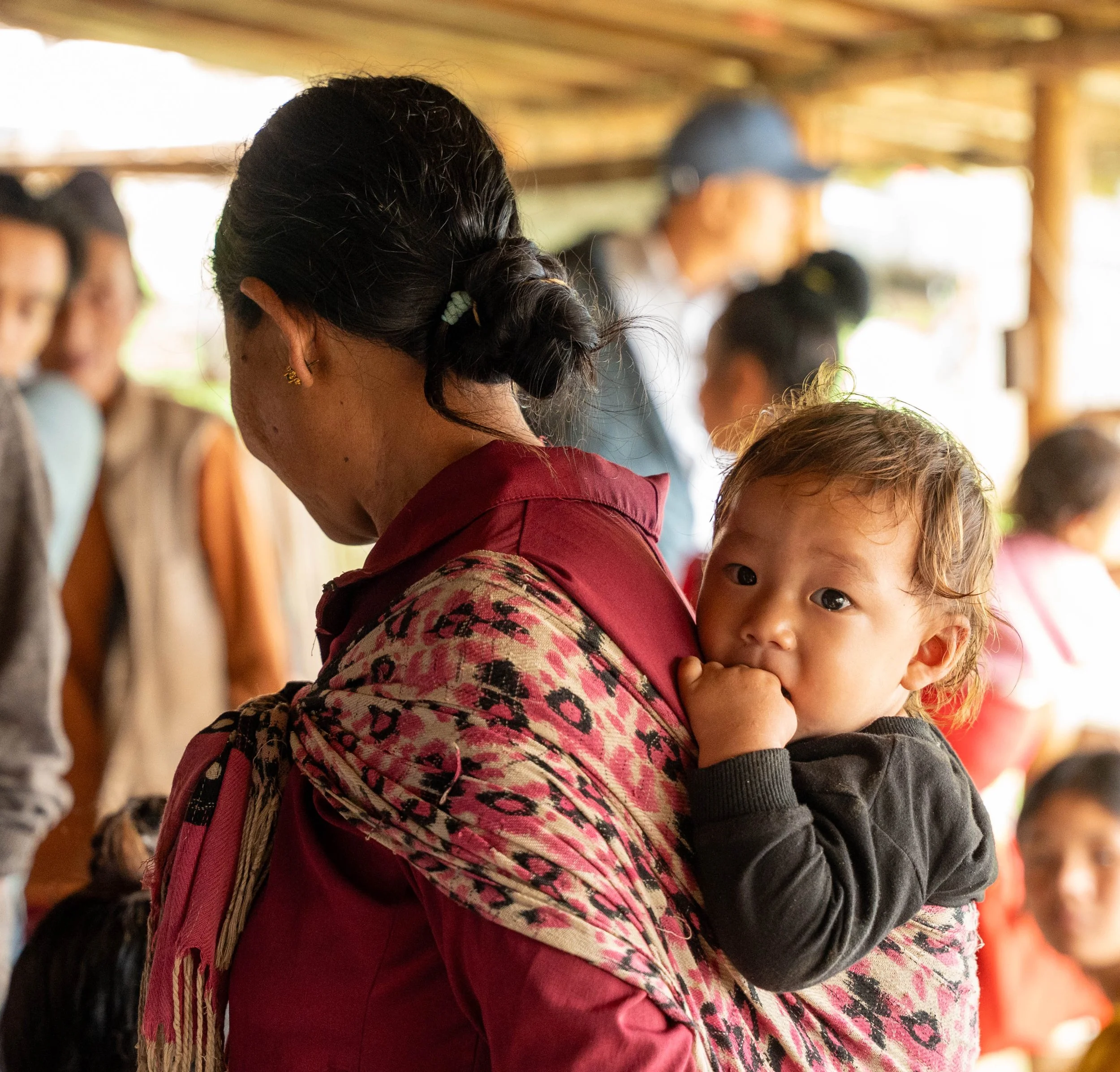 Close-up of a woman with black hair in a bun, wearing a patterned scarf and earrings, holding a young child with curly hair and dark eyes, in a social setting with other blurred people in the background.