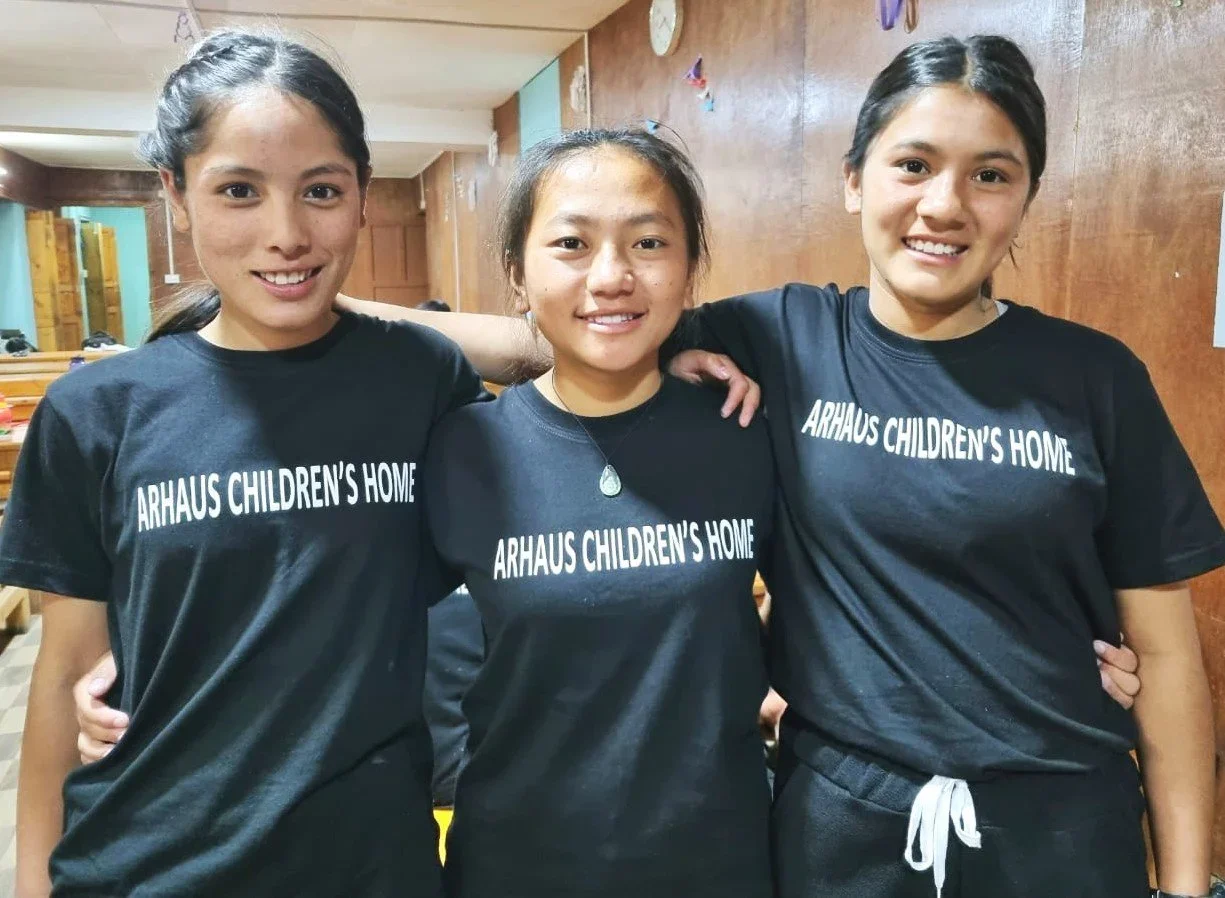 Three young women standing together in a room, wearing matching black t-shirts that say 'ARHAUS CHILDREN'S HOME'. They are smiling and have their arms around each other.