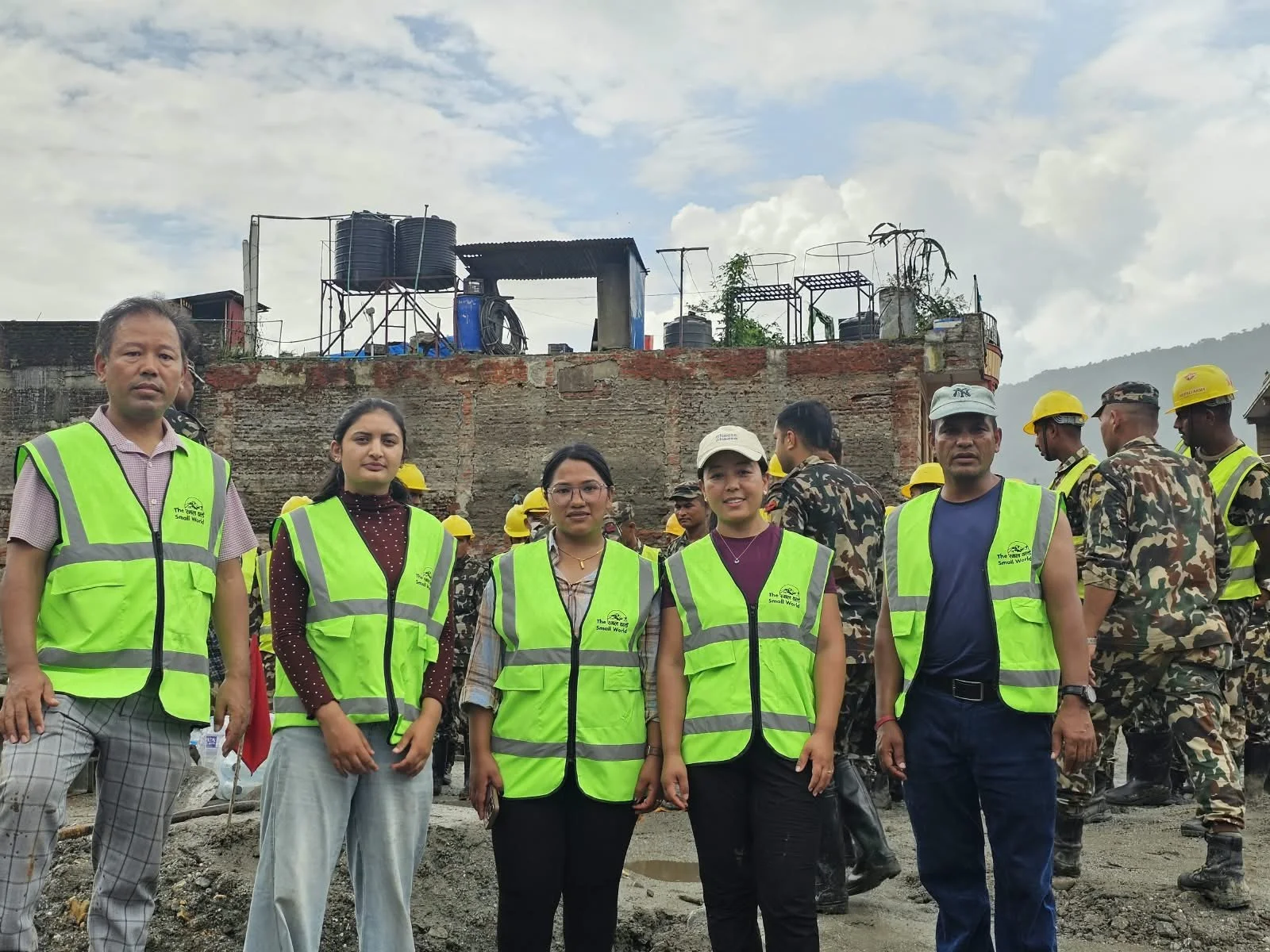 Group of five people wearing bright yellow safety vests standing in front of a construction site with workers and a brick wall.