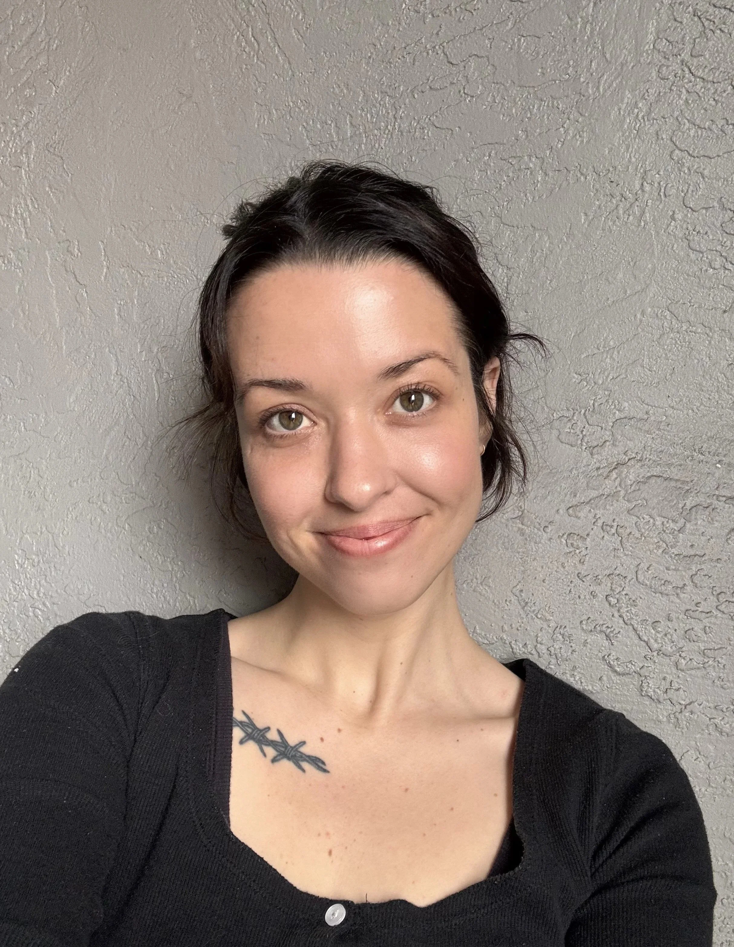 A woman with short dark hair and fair skin smiling at the camera, wearing a black top with a tattoo of three branches on her left shoulder against a textured wall.
