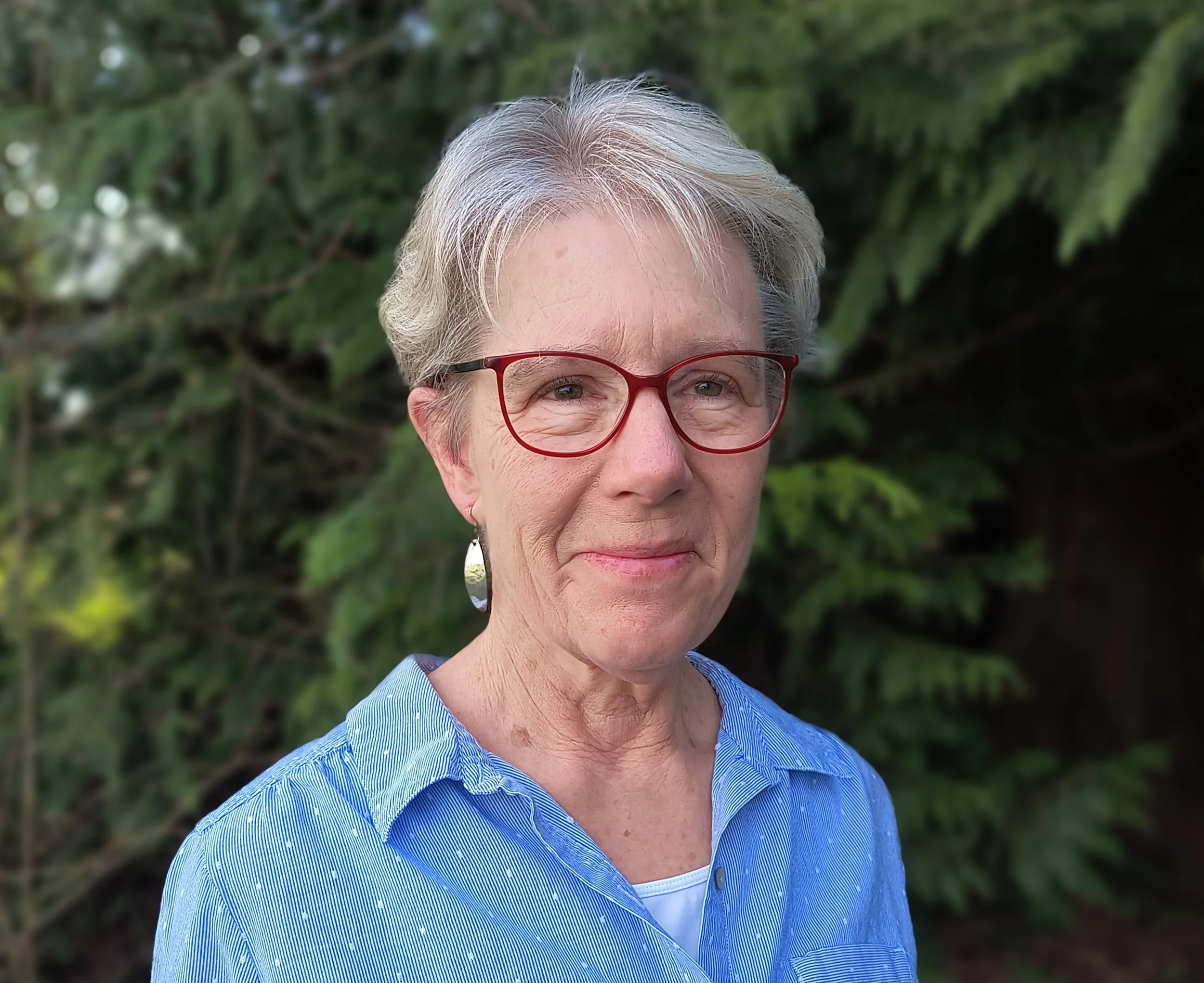 A woman wearing a purple hat and red glasses taking a selfie outdoors near a body of water with trees in the background.