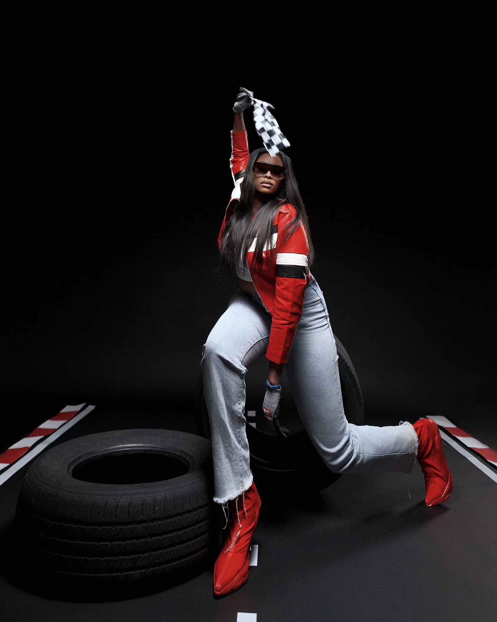 Woman in racing attire holding a checkered flag, posing among tires against a black background.
