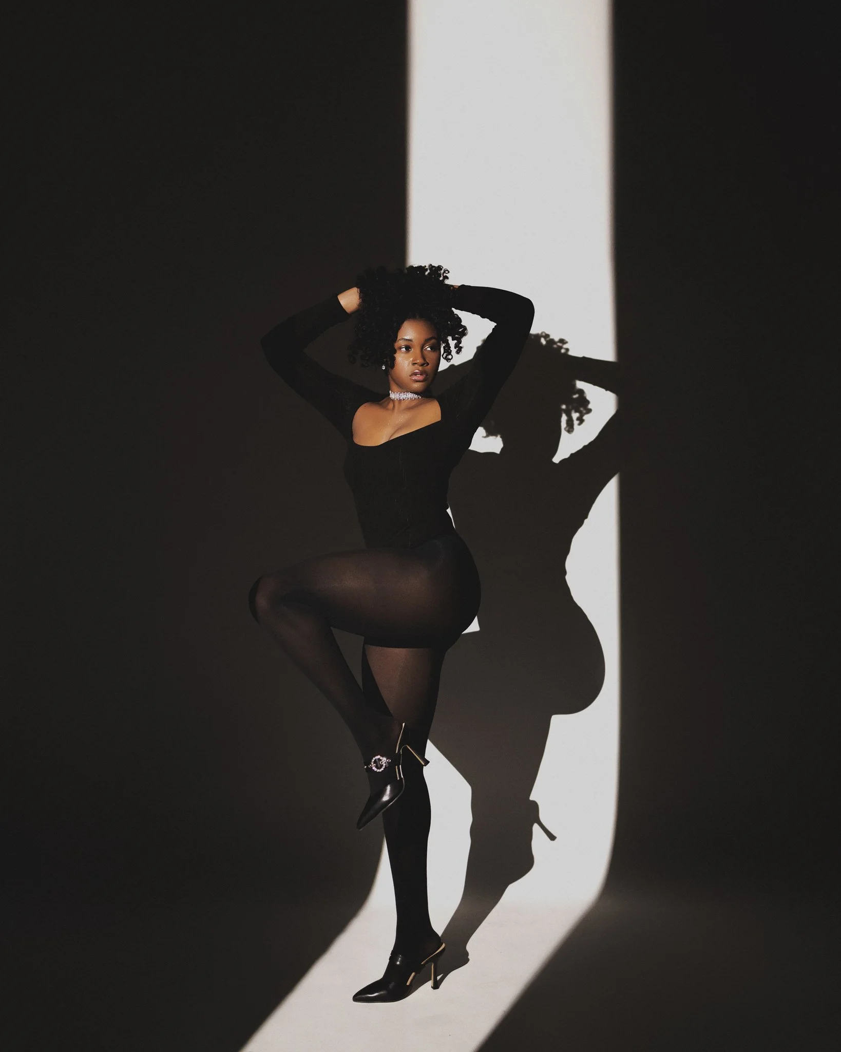 Woman with curly hair wears black dress and high heels, standing against a black and white background, casting a shadow.