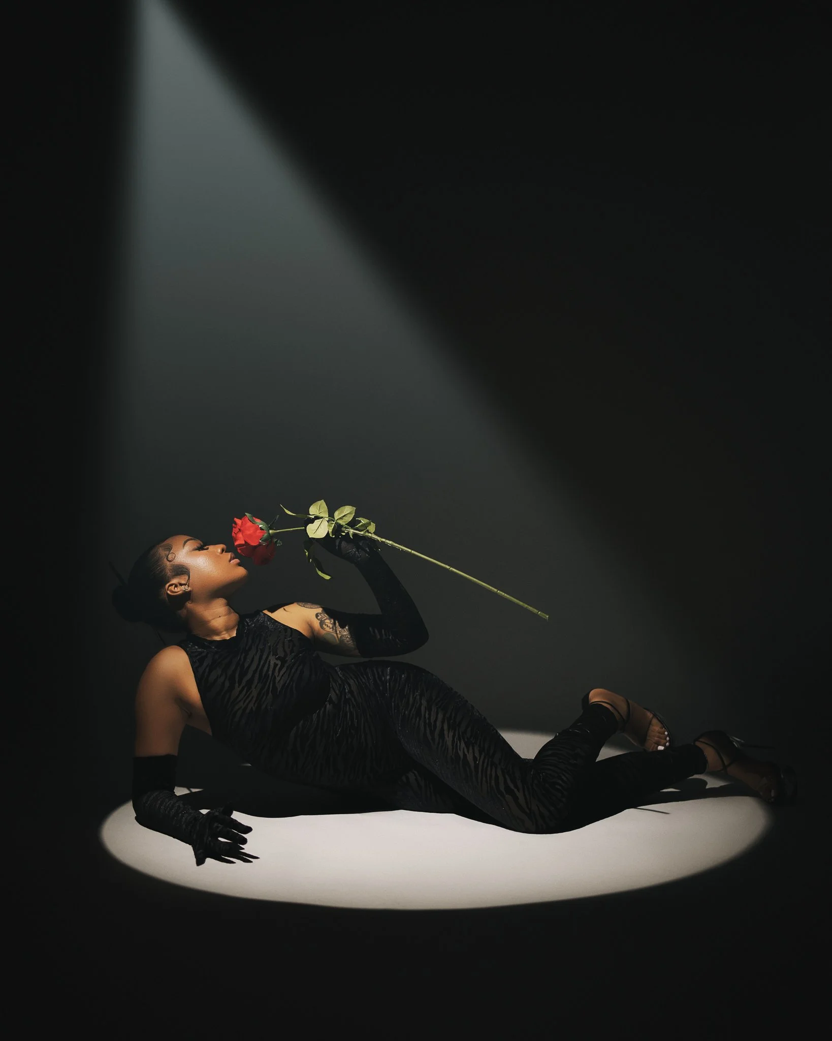 A woman lying on the floor in a spotlight, wearing a black dress and gloves, holding a red rose near her face, with dark background.