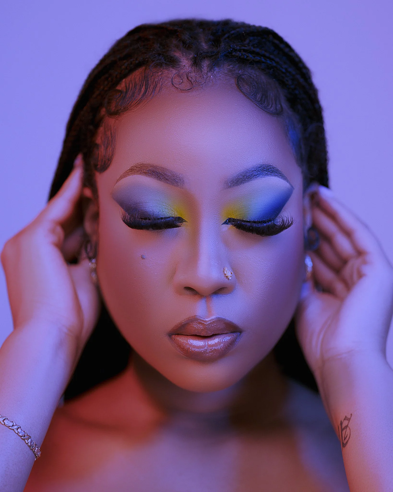 A woman with braided hair and makeup, eyes closed, touching her ears with both hands.