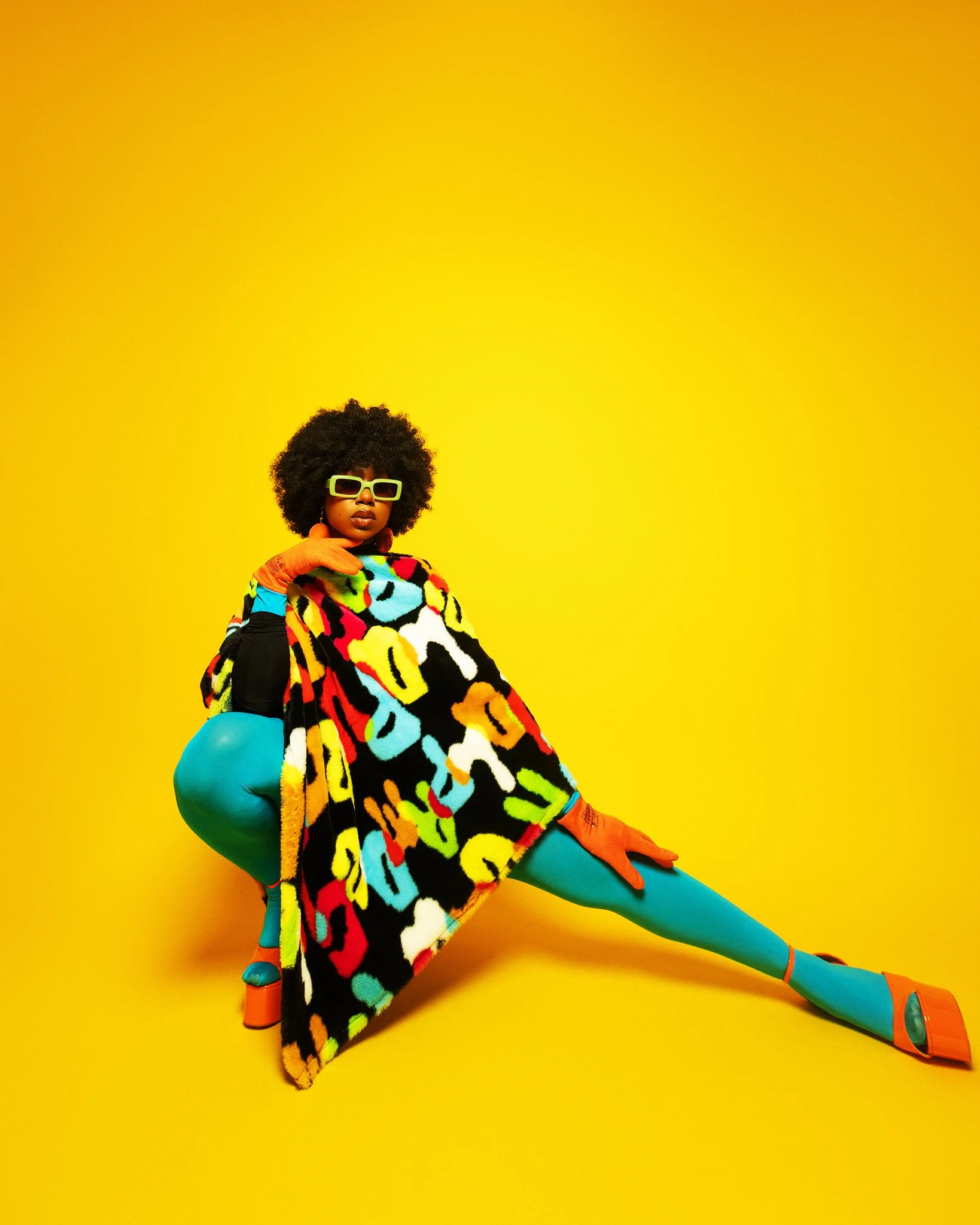 Woman with curly hair wearing colorful fashion accessories, sunglasses, and bright patterned clothing, striking a pose against a vibrant yellow background.