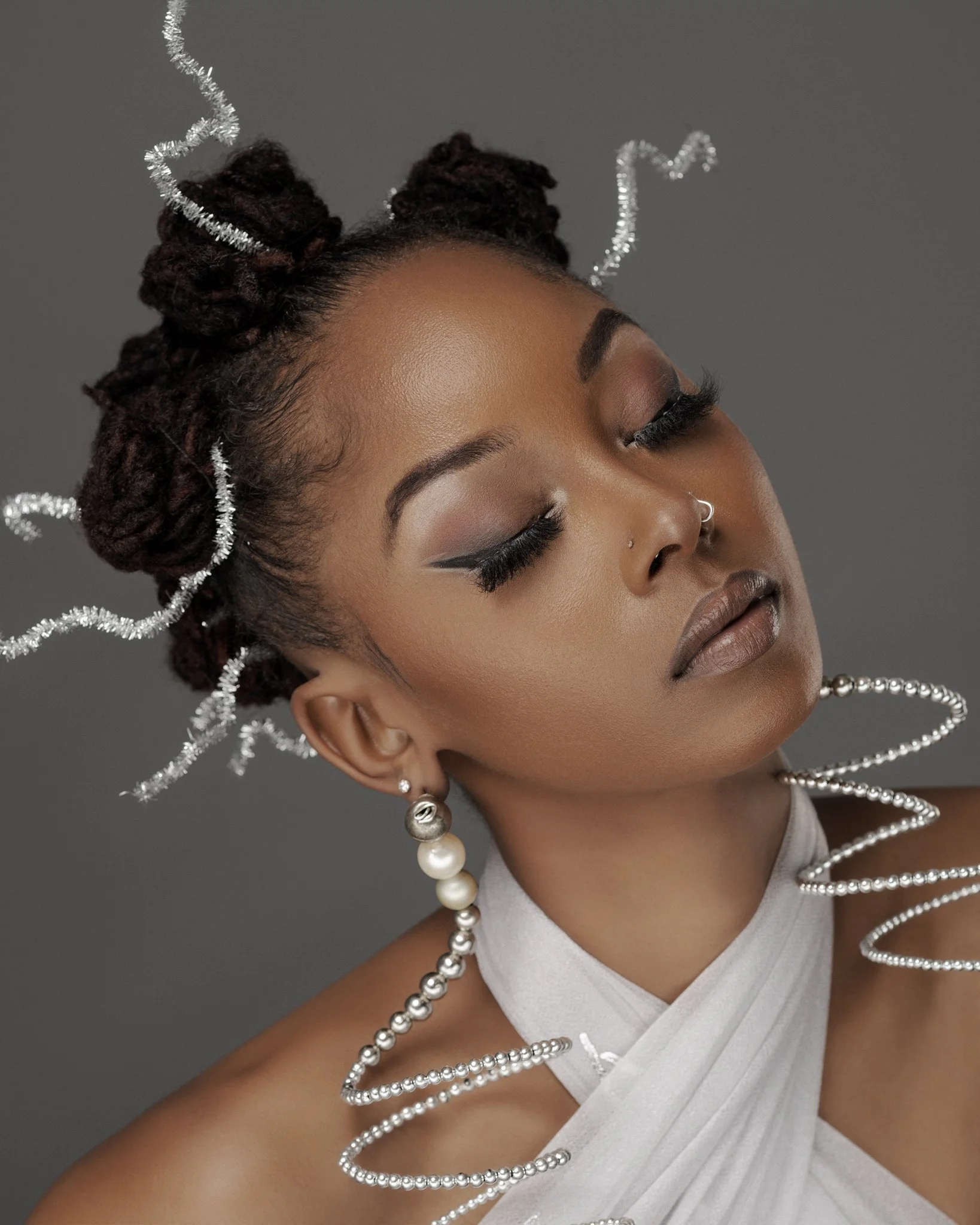 A woman with closed eyes, wearing makeup, earrings, and an outfit decorated with silver spirals and beads, against a gray background.