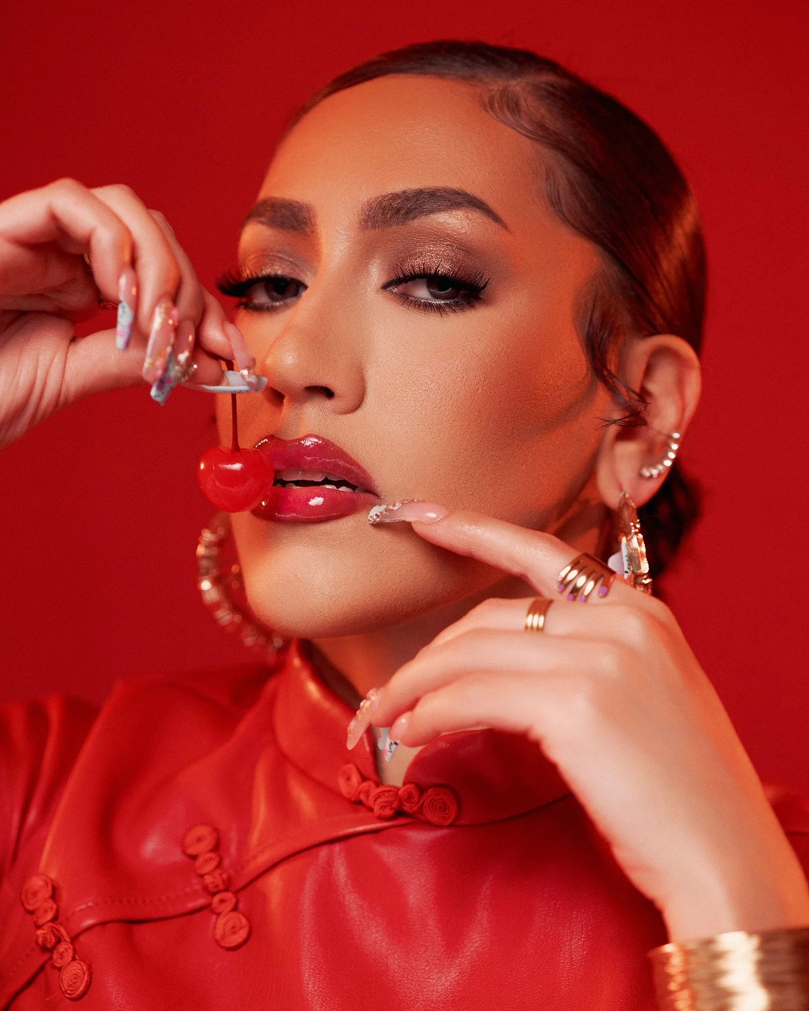 A woman with makeup and jewelry holding a cherry near her lips against a red background.