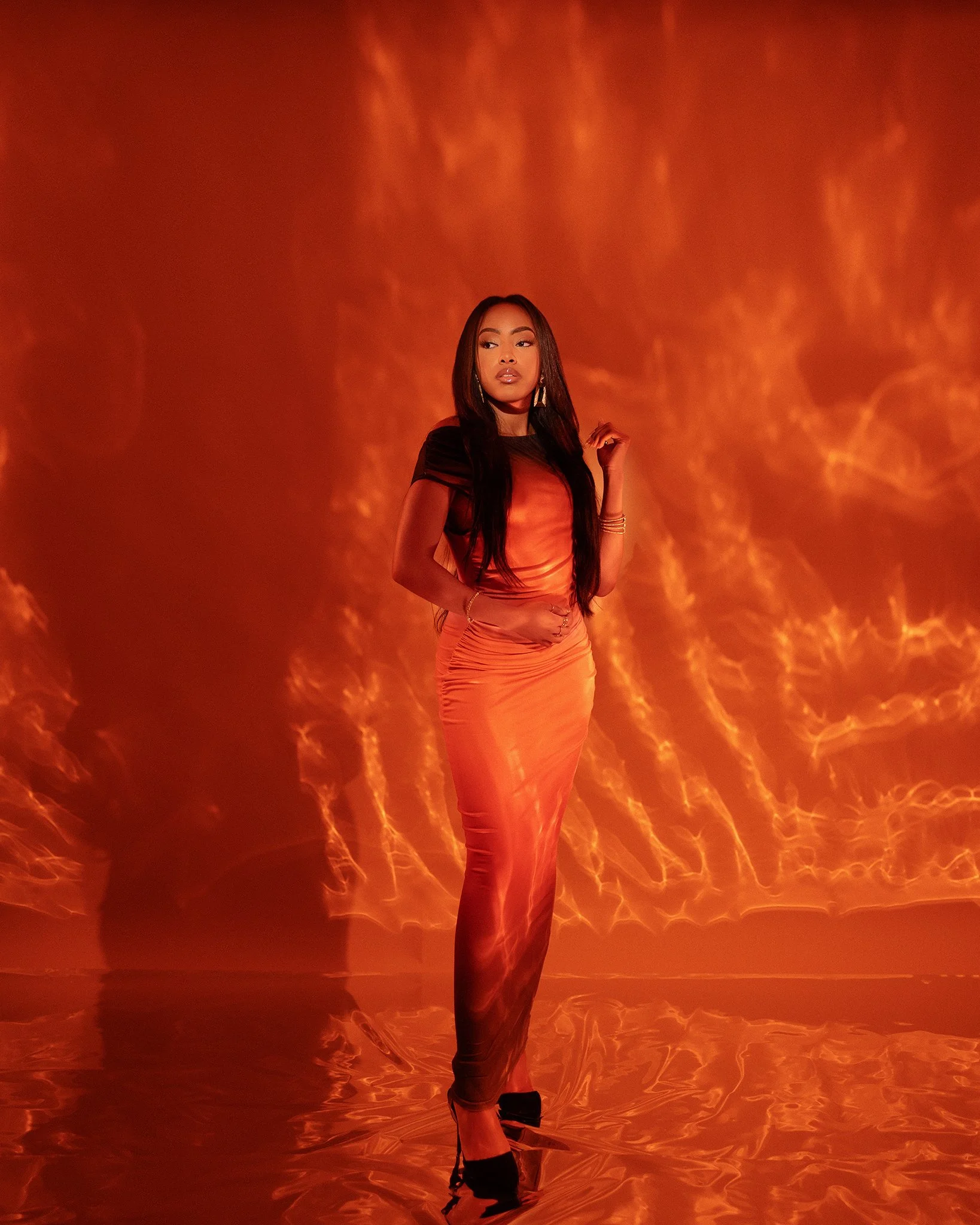 A woman in a shiny orange dress and black high heels standing in front of a fiery orange and red background.