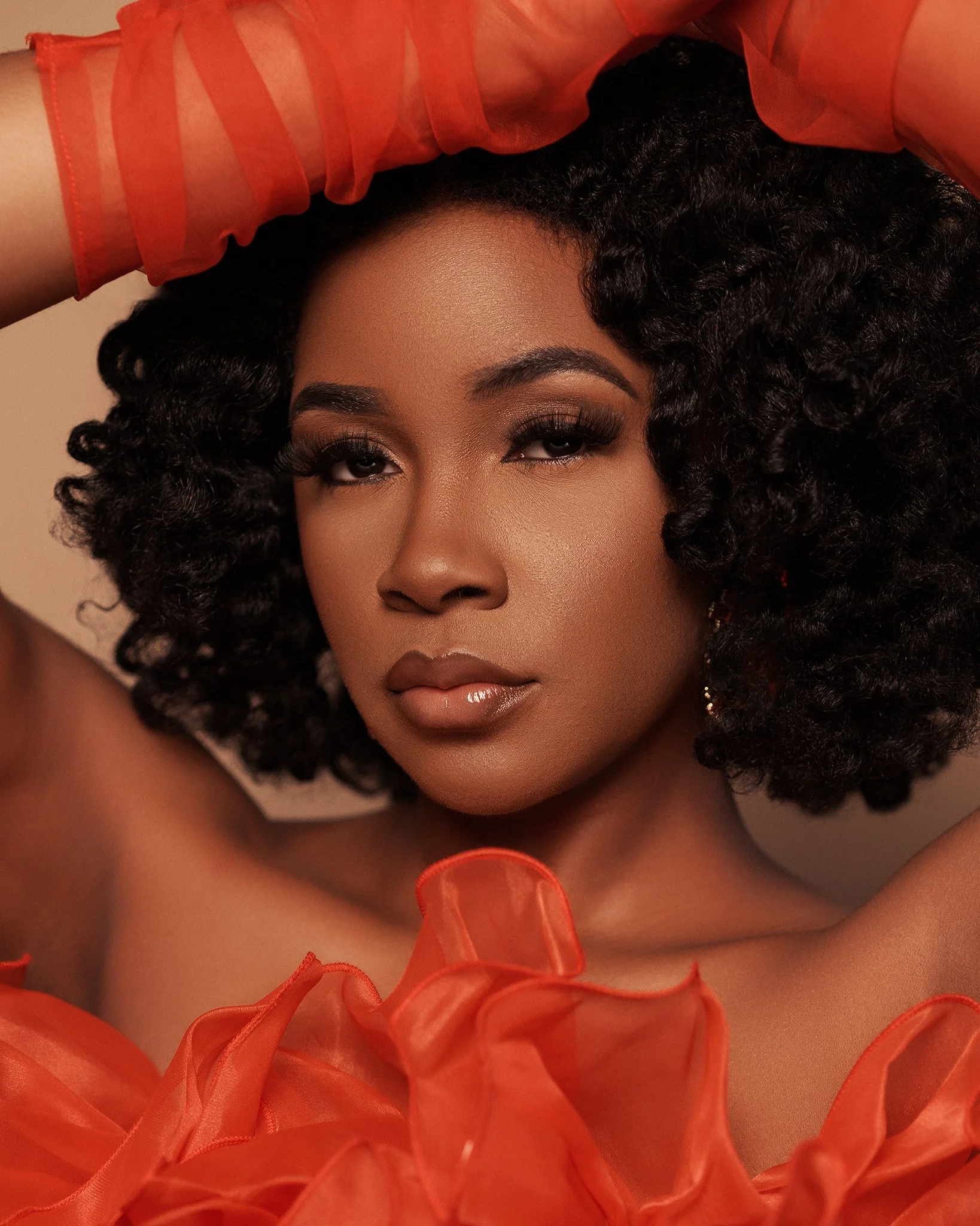 A woman with curly black hair, wearing sheer orange fabric, posing with her arms raised above her head.