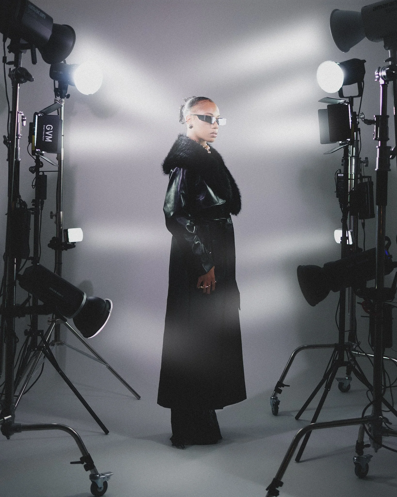 A woman posing in a photography studio with professional lighting, wearing dark fashion accessories including sunglasses, a fur-collared leather jacket, and long black skirt.